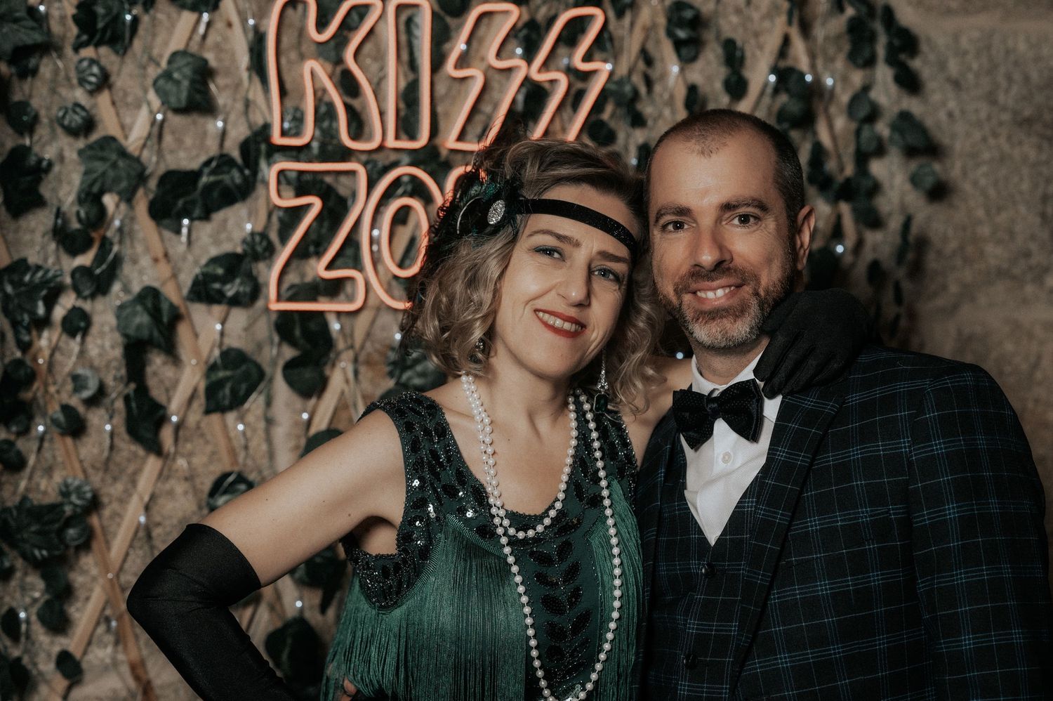A couple poses together in front of a textured wall while wearing beaded necklaces at what appears to be a party.