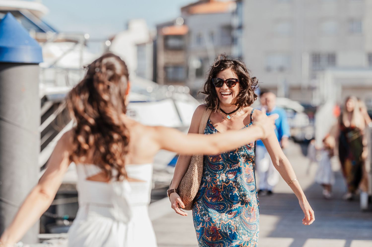 People enjoying a sunny day walking on a marina boardwalk with boats and buildings in the background.