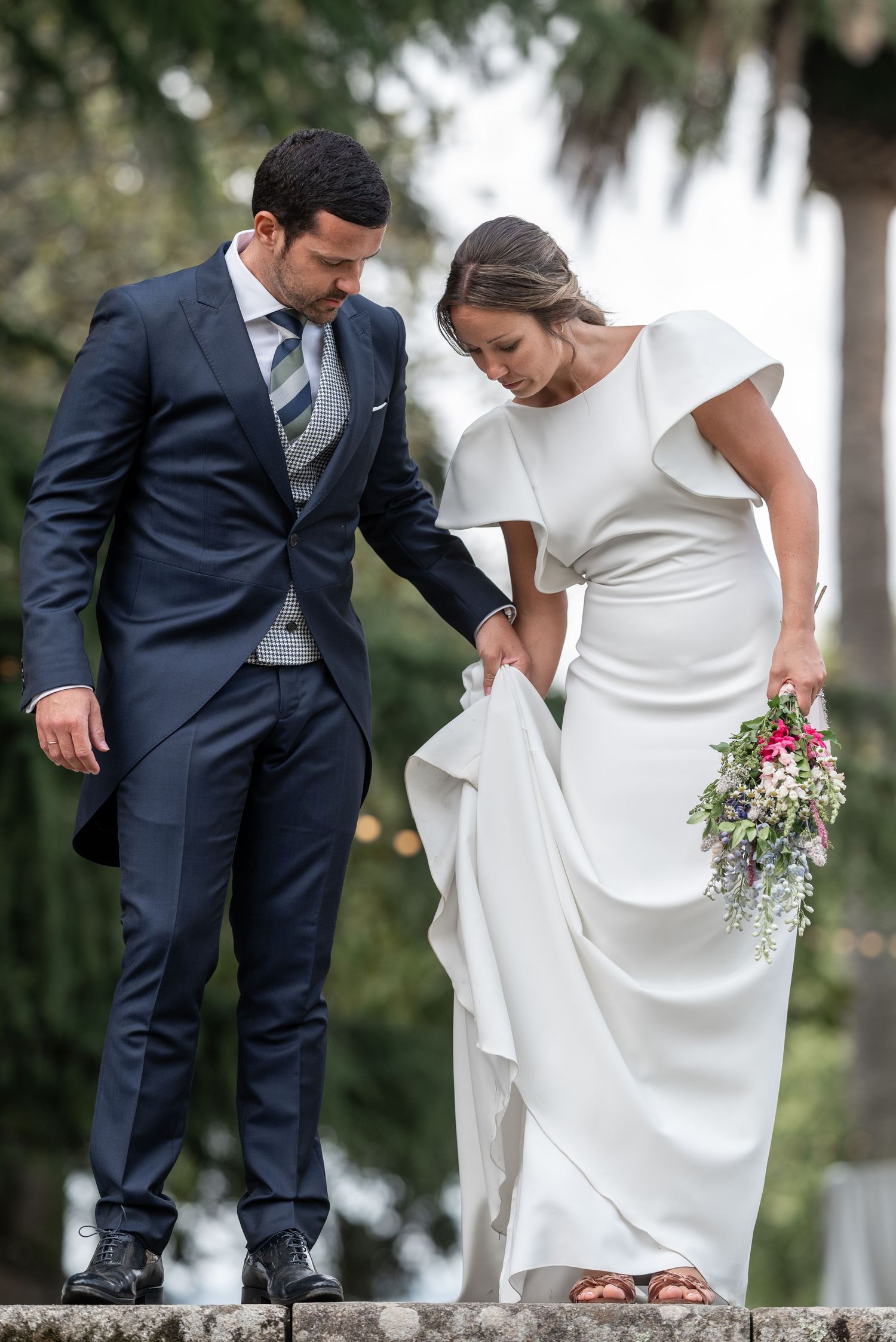 A couple in wedding attire shares a tender moment outdoors as the groom helps adjust the bride's white dress.