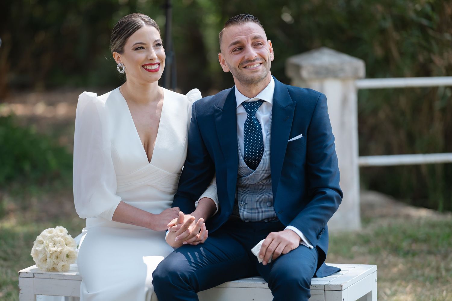 A bride and groom share a tender moment outdoors while seated together on their wedding day.