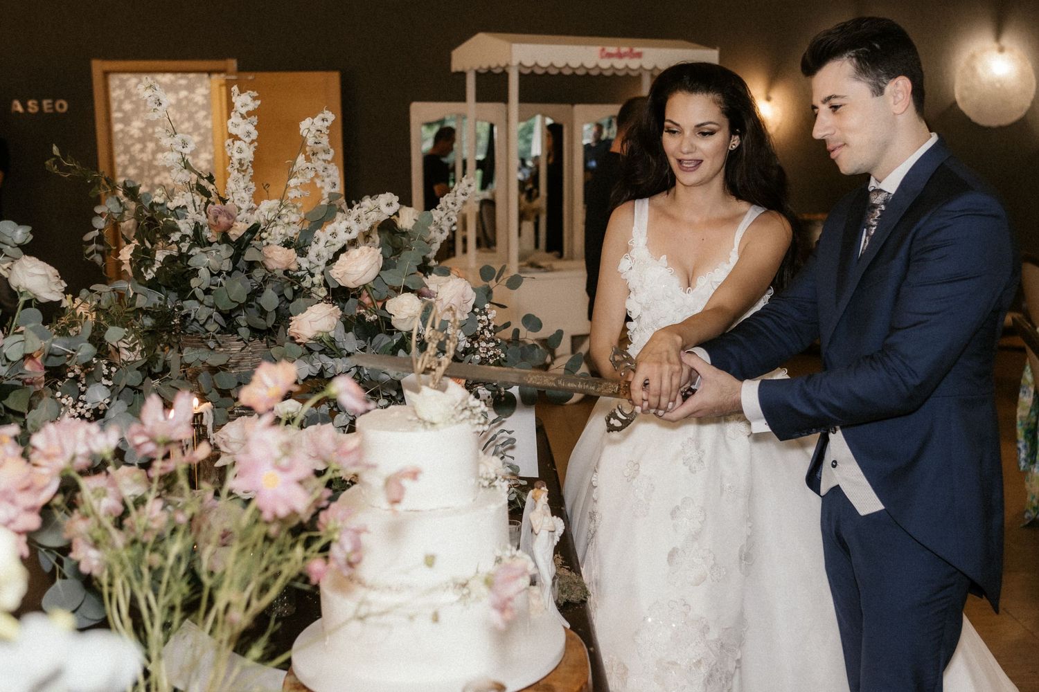 A couple shares an intimate moment cutting their wedding cake together at an elegant reception decorated with floral arrangements.