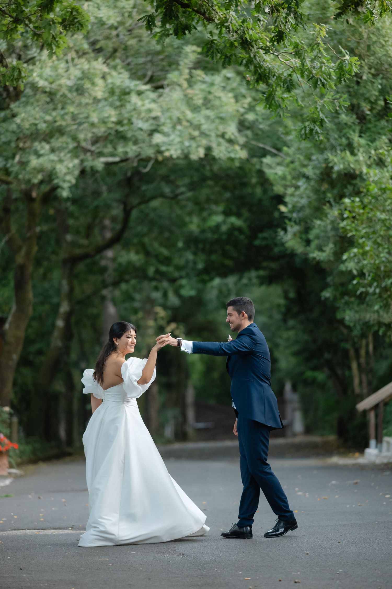 A bride and groom dance together on a tree-lined path during their wedding photoshoot.