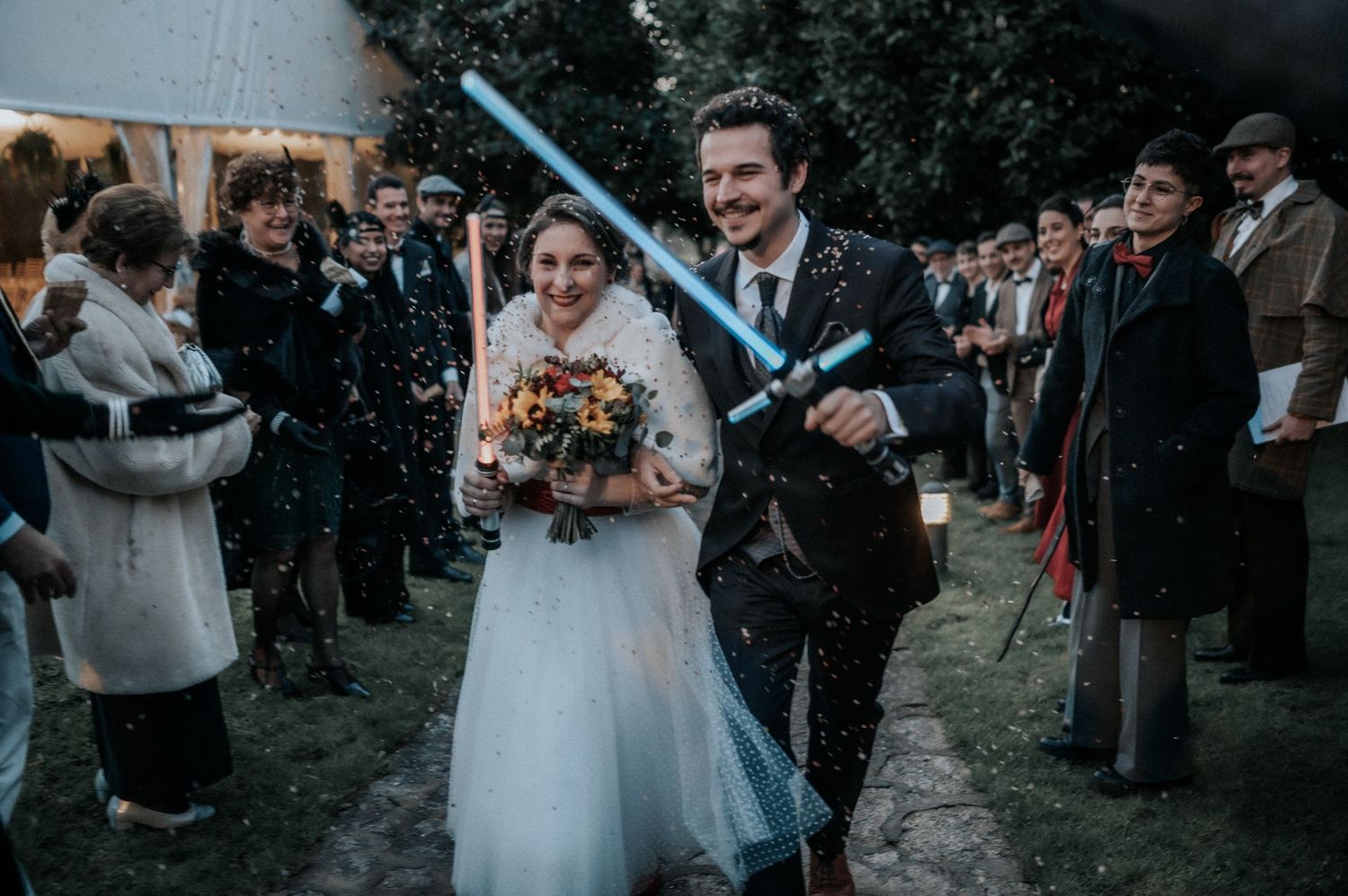 Wedding party members hold glowing blue lightsabers in the air during an outdoor nighttime ceremony.