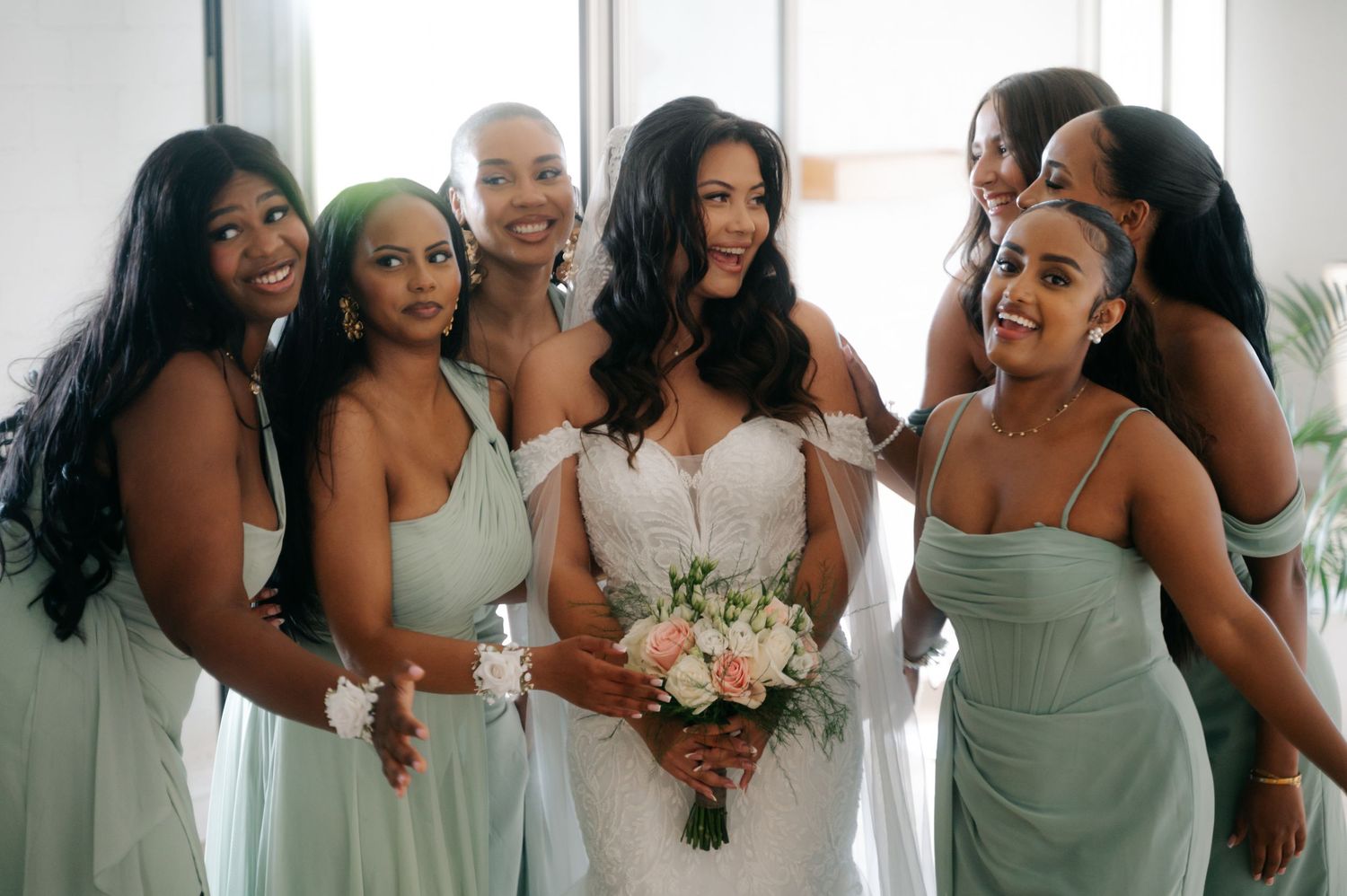 A bridal party in sage green dresses smiles together while holding a bouquet of blush and white flowers.