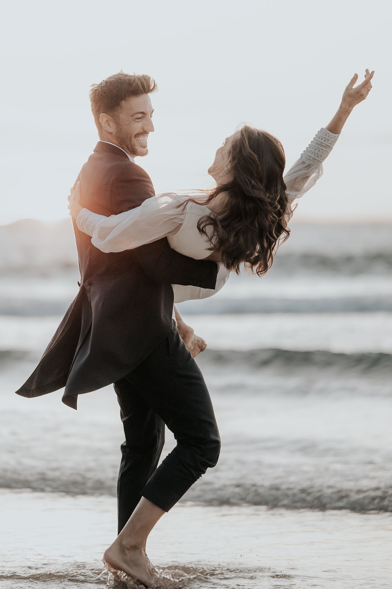 Playful couple dancing and twirling on beach at sunset during wedding photoshoot.