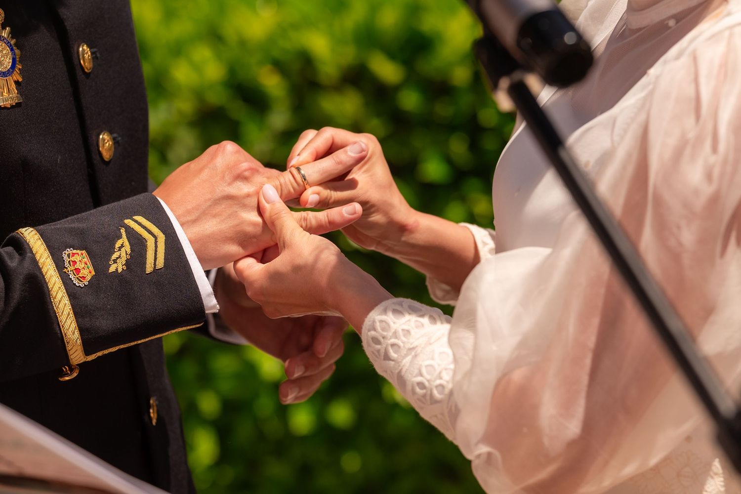 Close-up of hands exchanging wedding rings during an outdoor ceremony with green foliage in background.