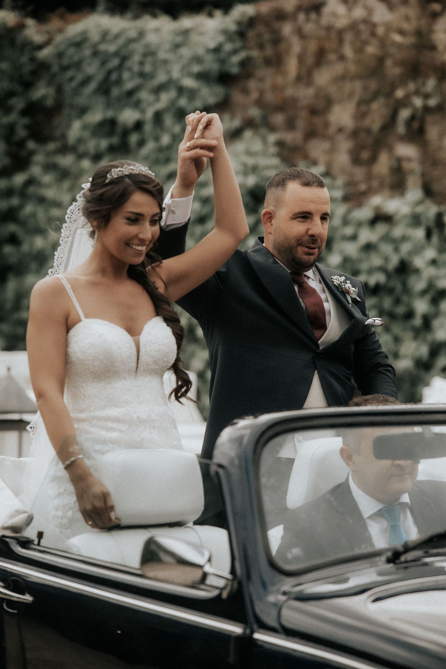 A couple in wedding attire wave while seated in a classic black convertible car during their celebration.
