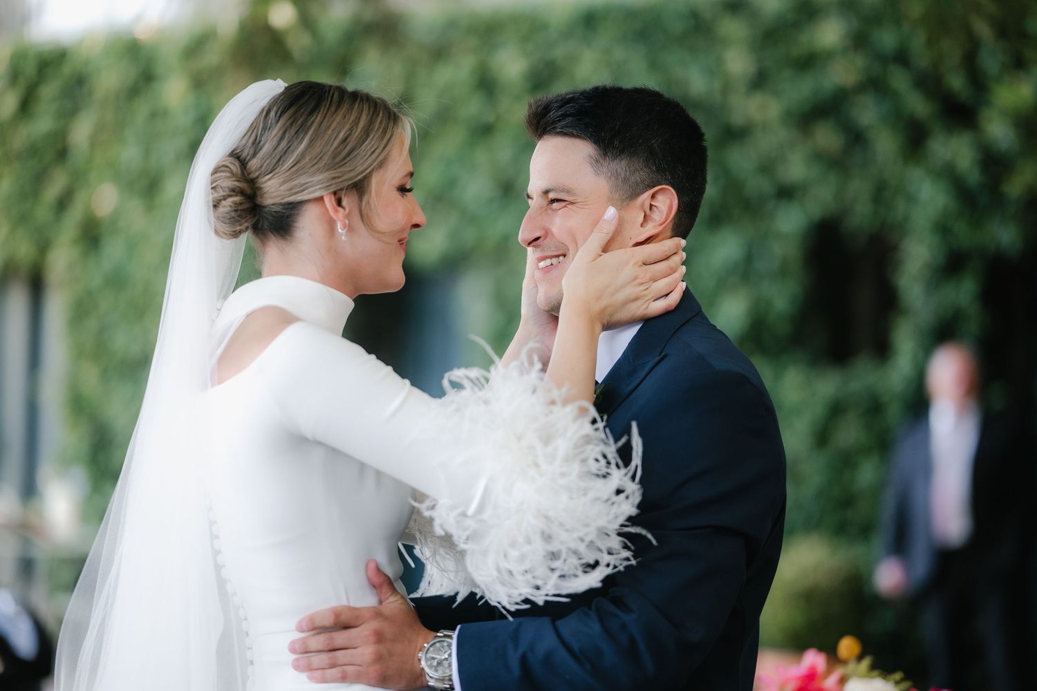 A tender moment between newlyweds outdoors with the bride wearing a white dress with feather details and groom in navy suit.