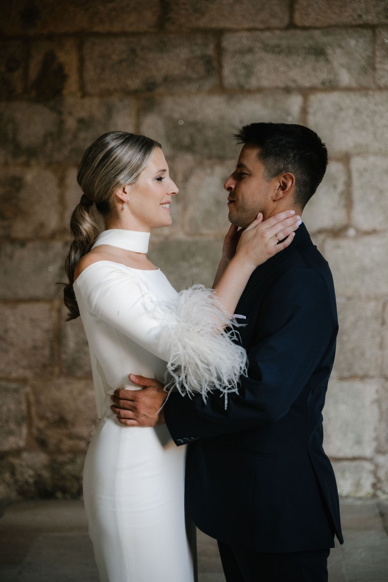 Tender moment between newlyweds against stone wall backdrop showing elegant white dress with feather details.