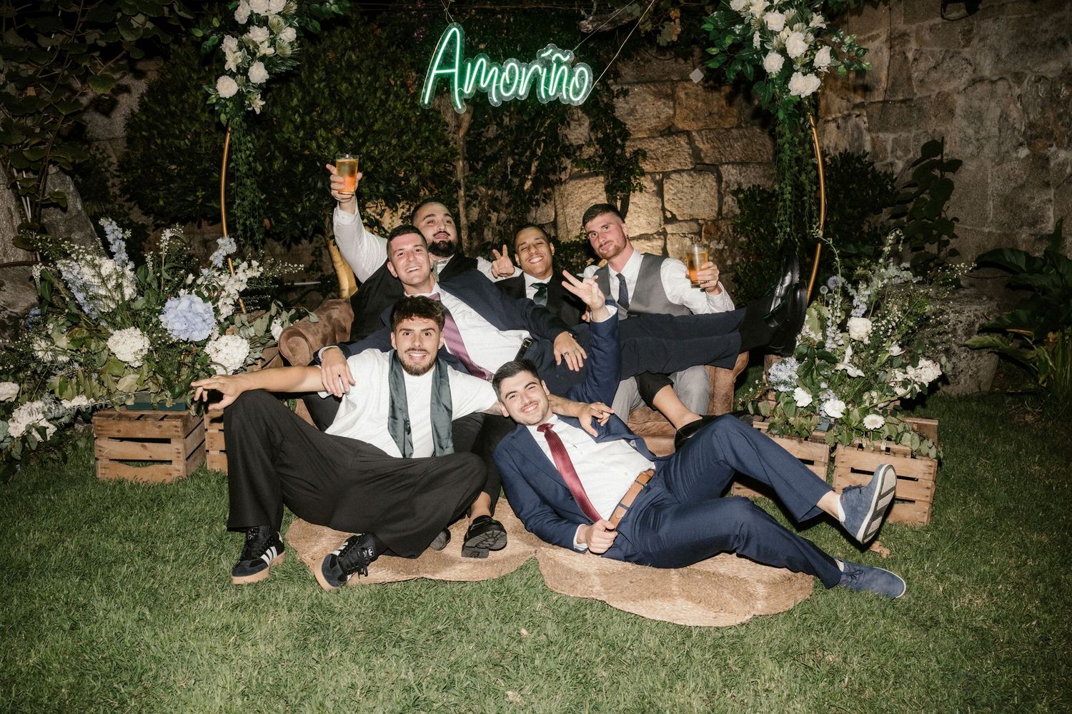 Wedding party poses playfully under a neon sign and floral arch at an outdoor venue.