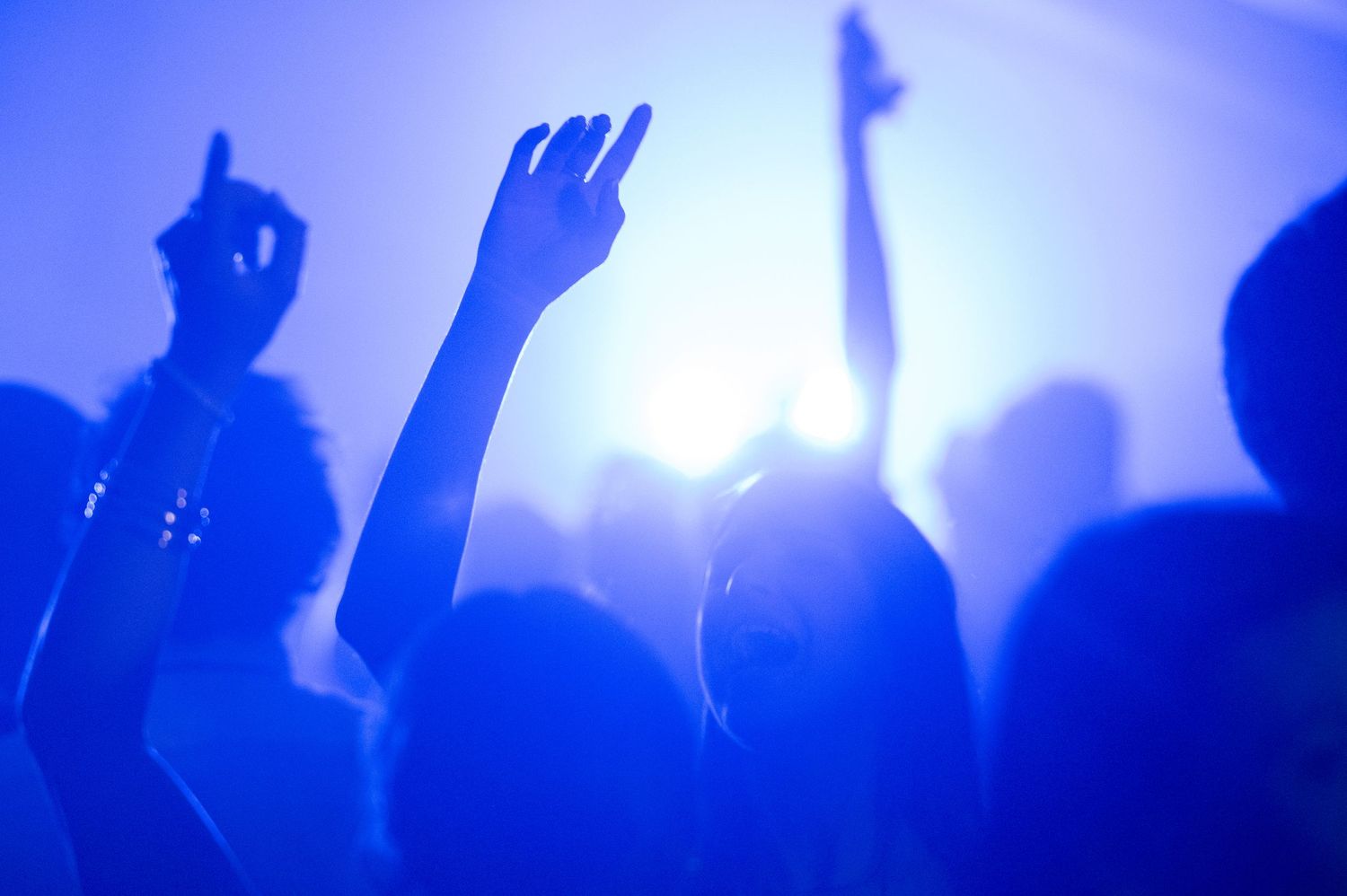 Silhouetted crowd raises hands at a concert with bright blue stage lighting and atmospheric effects.