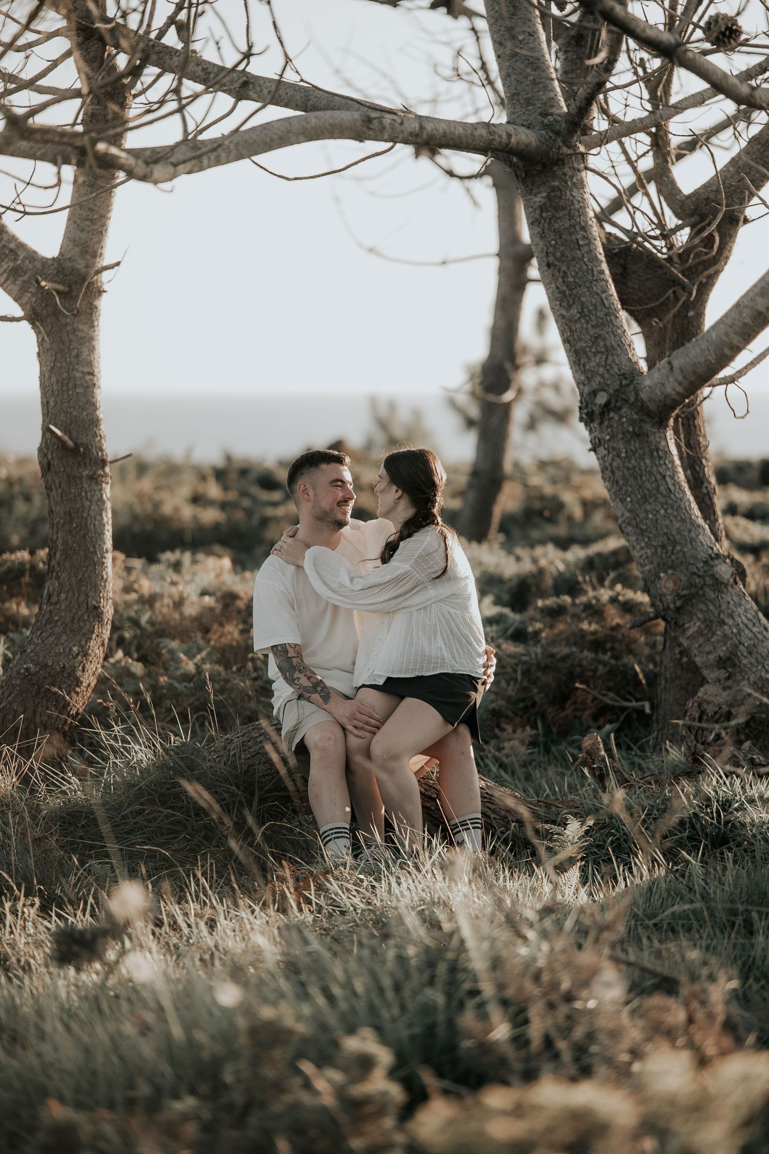 A romantic couple embraces under tall trees during a golden sunset in a natural outdoor setting.