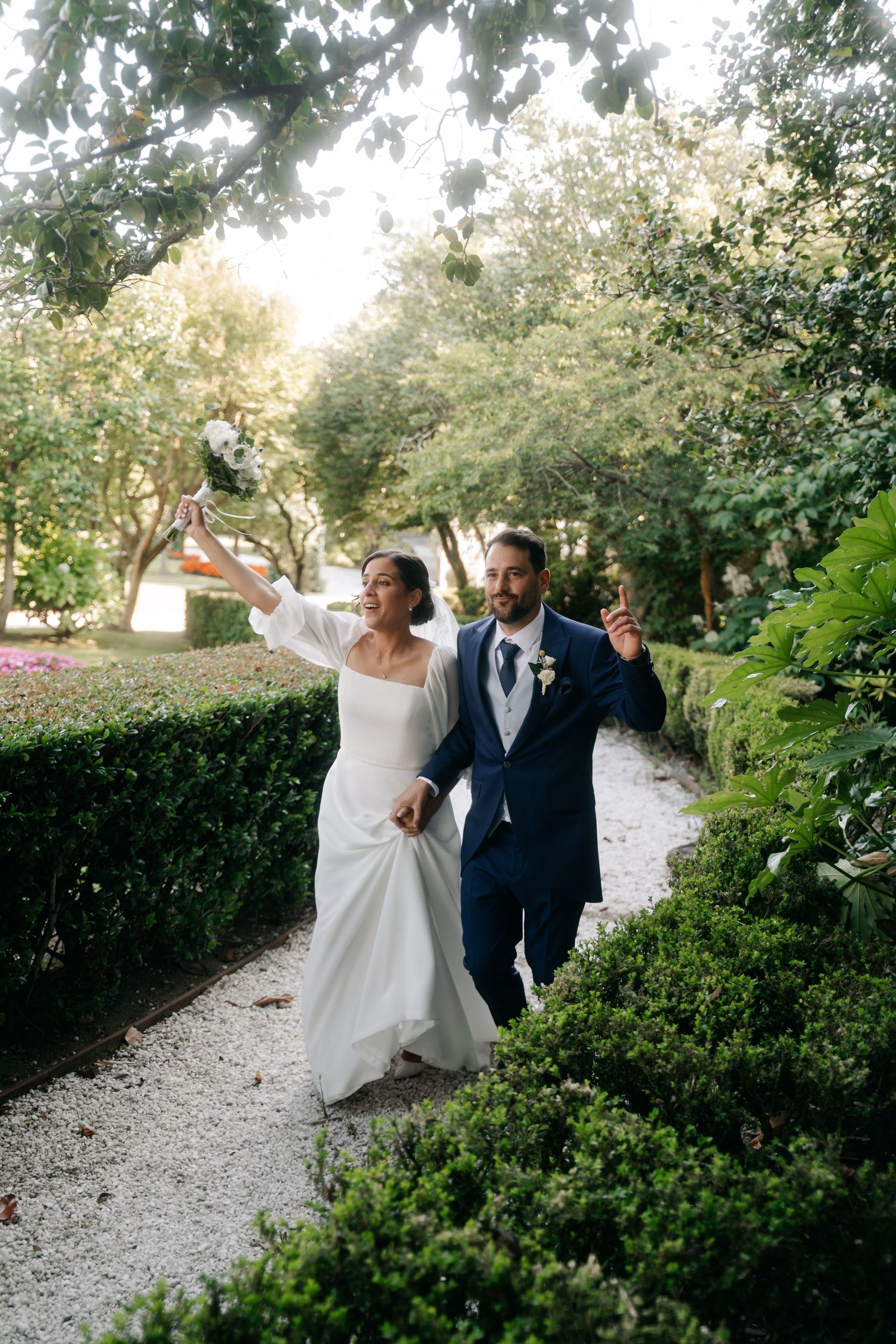 A joyful couple dances together on a gravel path surrounded by lush green garden hedges on their wedding day.