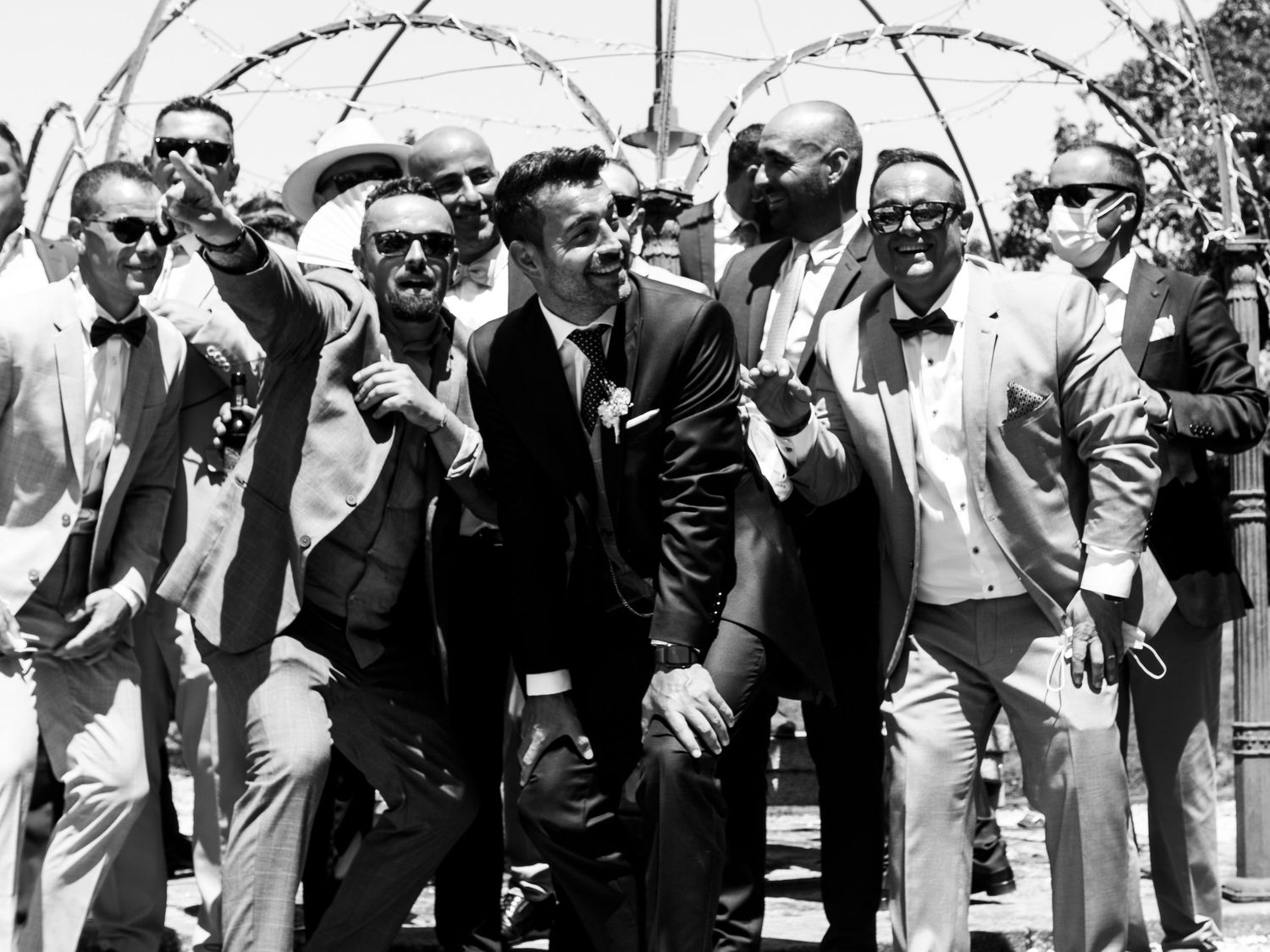 A black and white photo shows a large group of men in suits and sunglasses standing together outdoors.