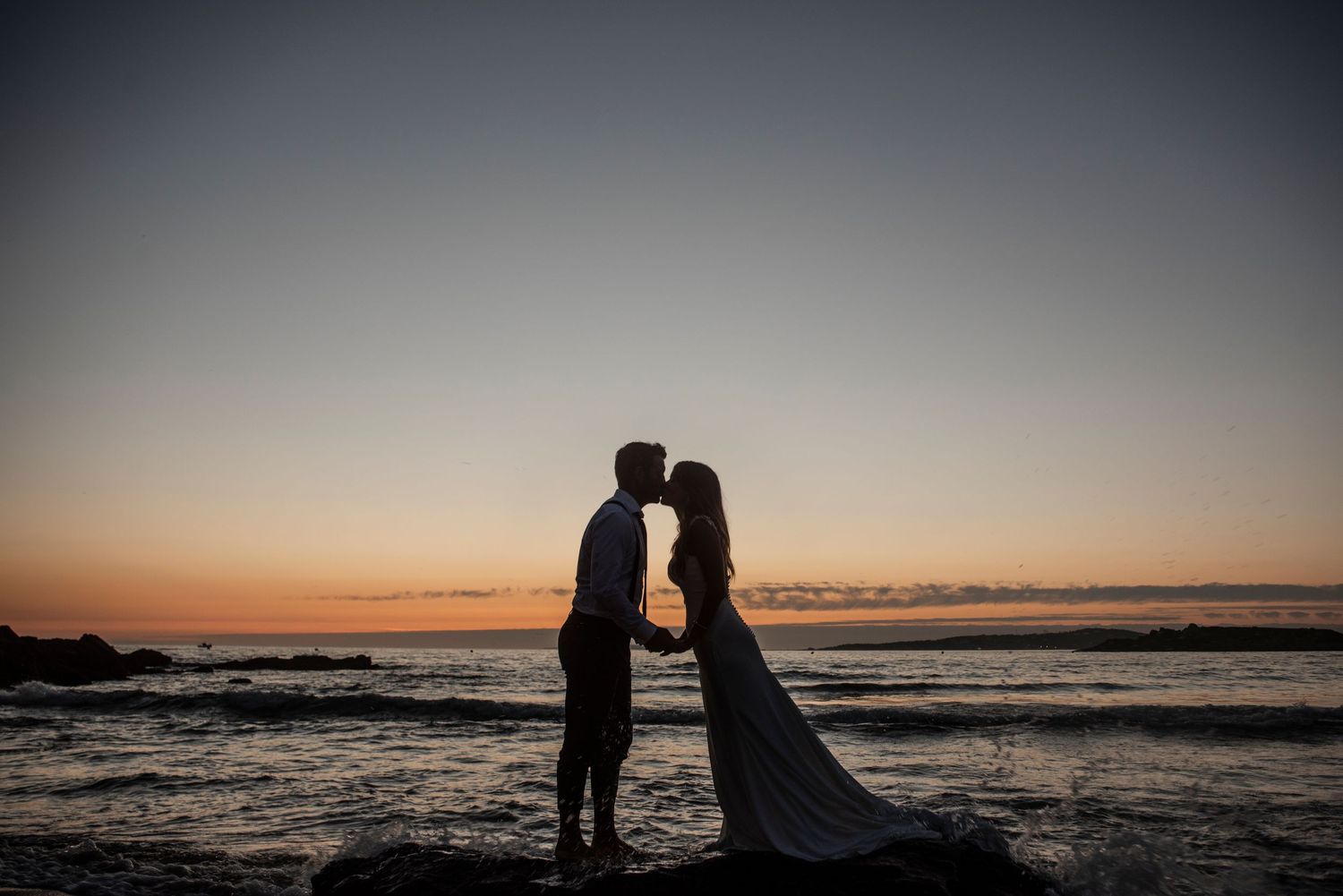 Silhouettes of a couple stand together against a colorful beach sunset horizon.