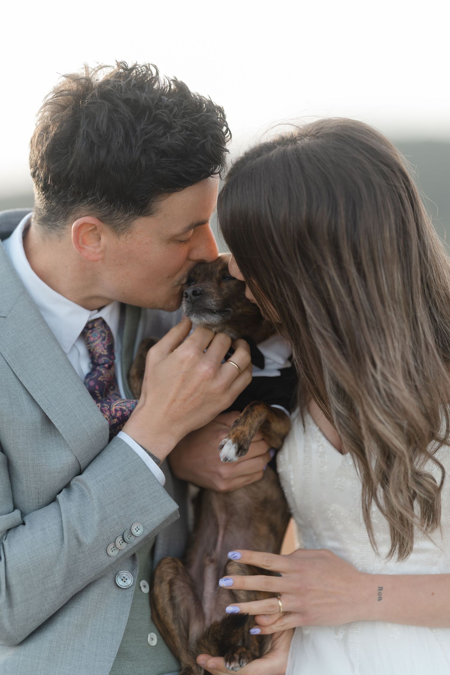 Bride and groom in wedding attire kissing their small brown dog together outdoors on their wedding day.