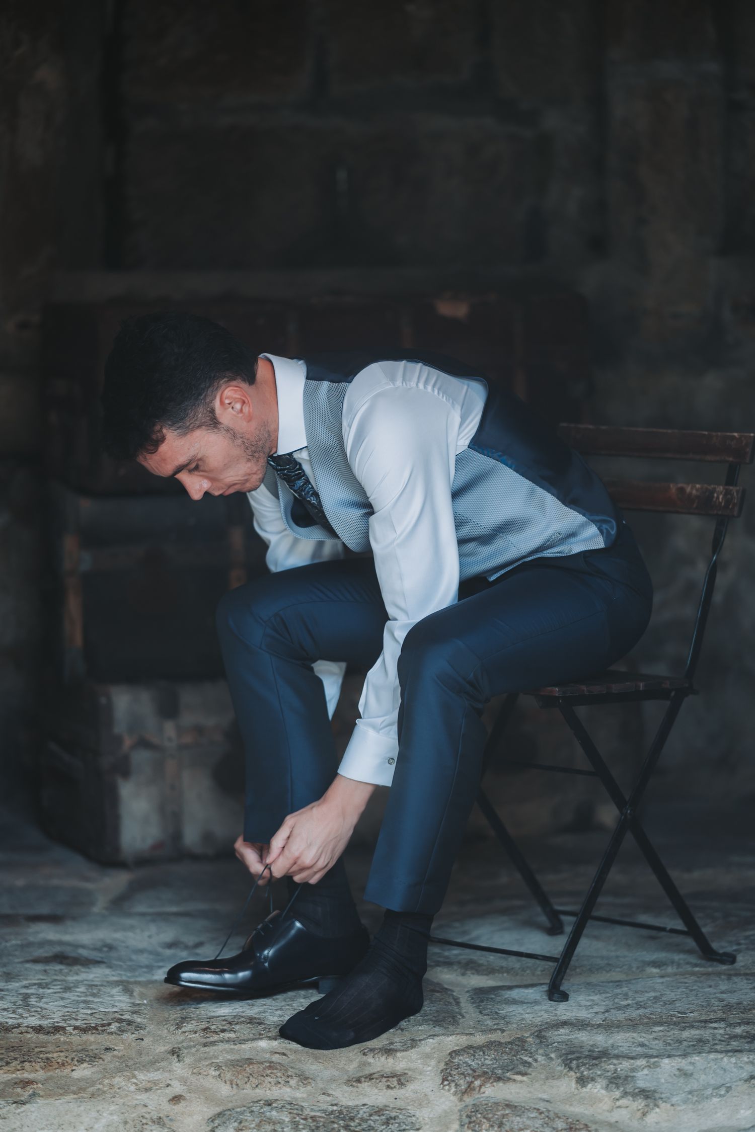 Person in formal attire sitting on dark chair tying black dress shoes.