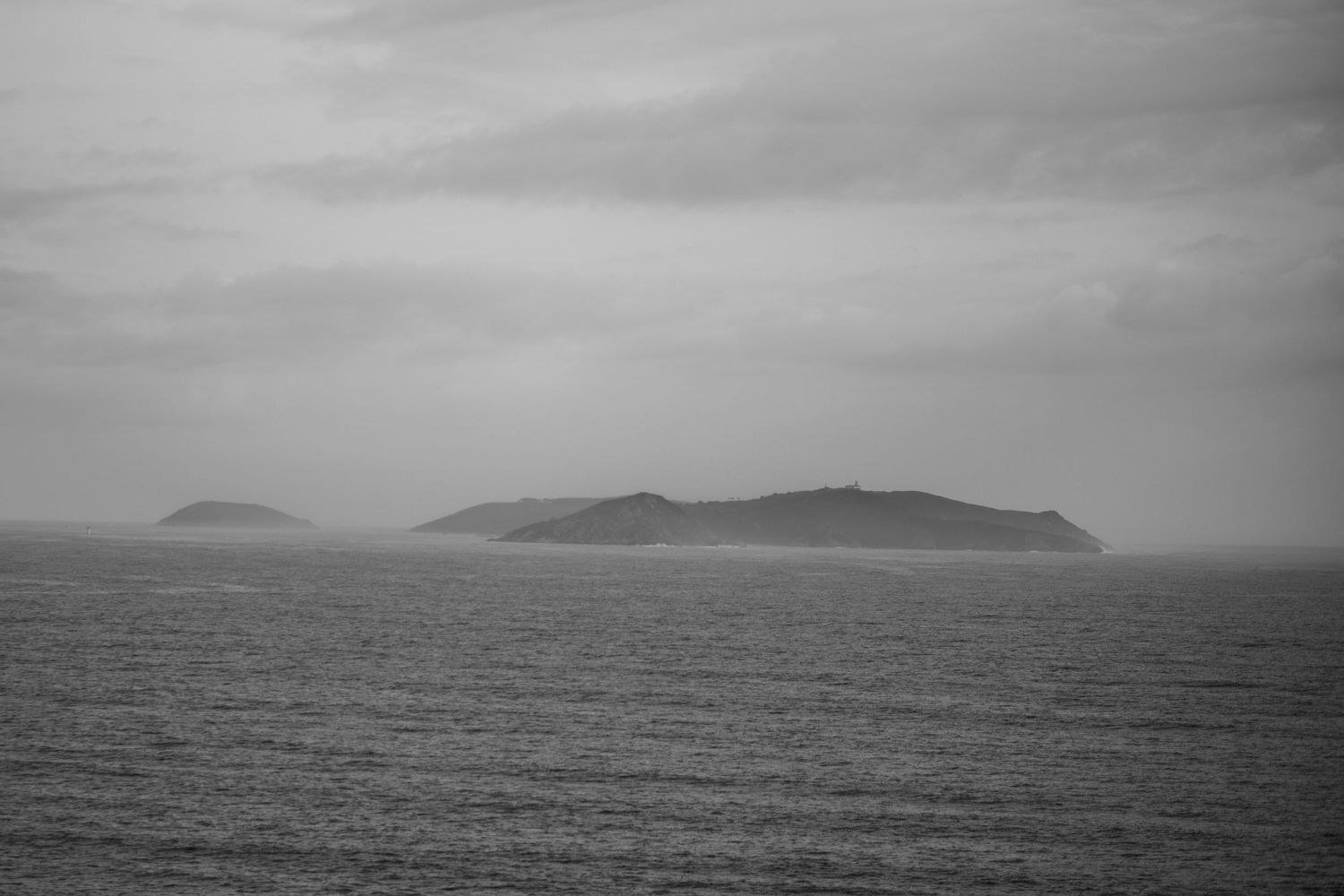 A misty black and white seascape shows distant mountainous islands emerging from the calm ocean waters.