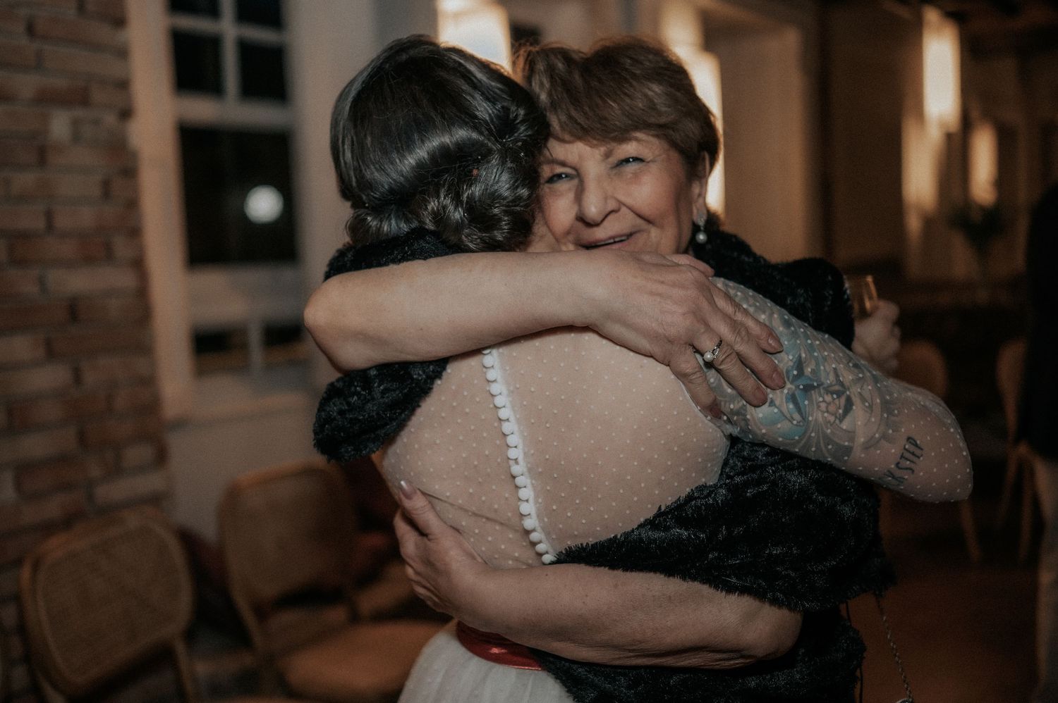 Two people share a warm embrace during an intimate indoor celebration with soft lighting and brick walls.