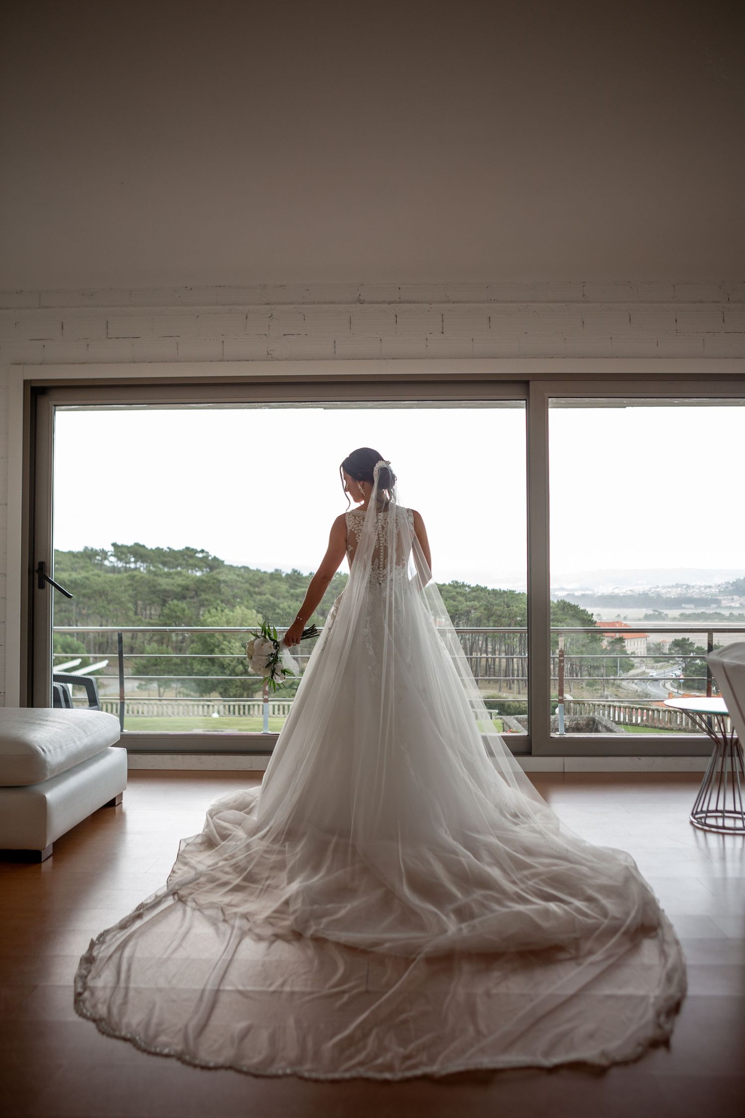 A dramatic full-length view of a bride's wedding dress and veil trailing across a modern room with panoramic windows.