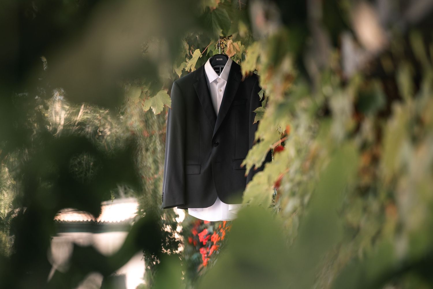 A couple in formal wedding attire stands in a lush garden setting with sunlight filtering through tree branches.