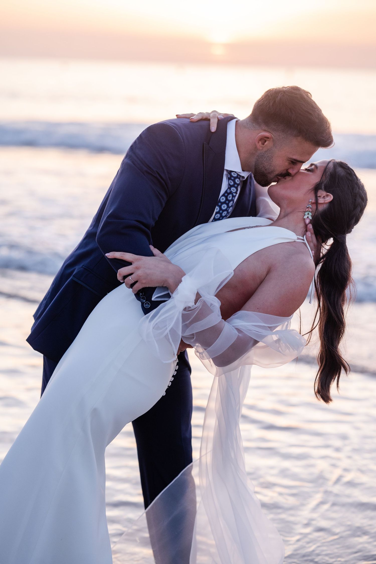 A romantic beach wedding couple shares a passionate kiss at sunset with ocean waves in the background.