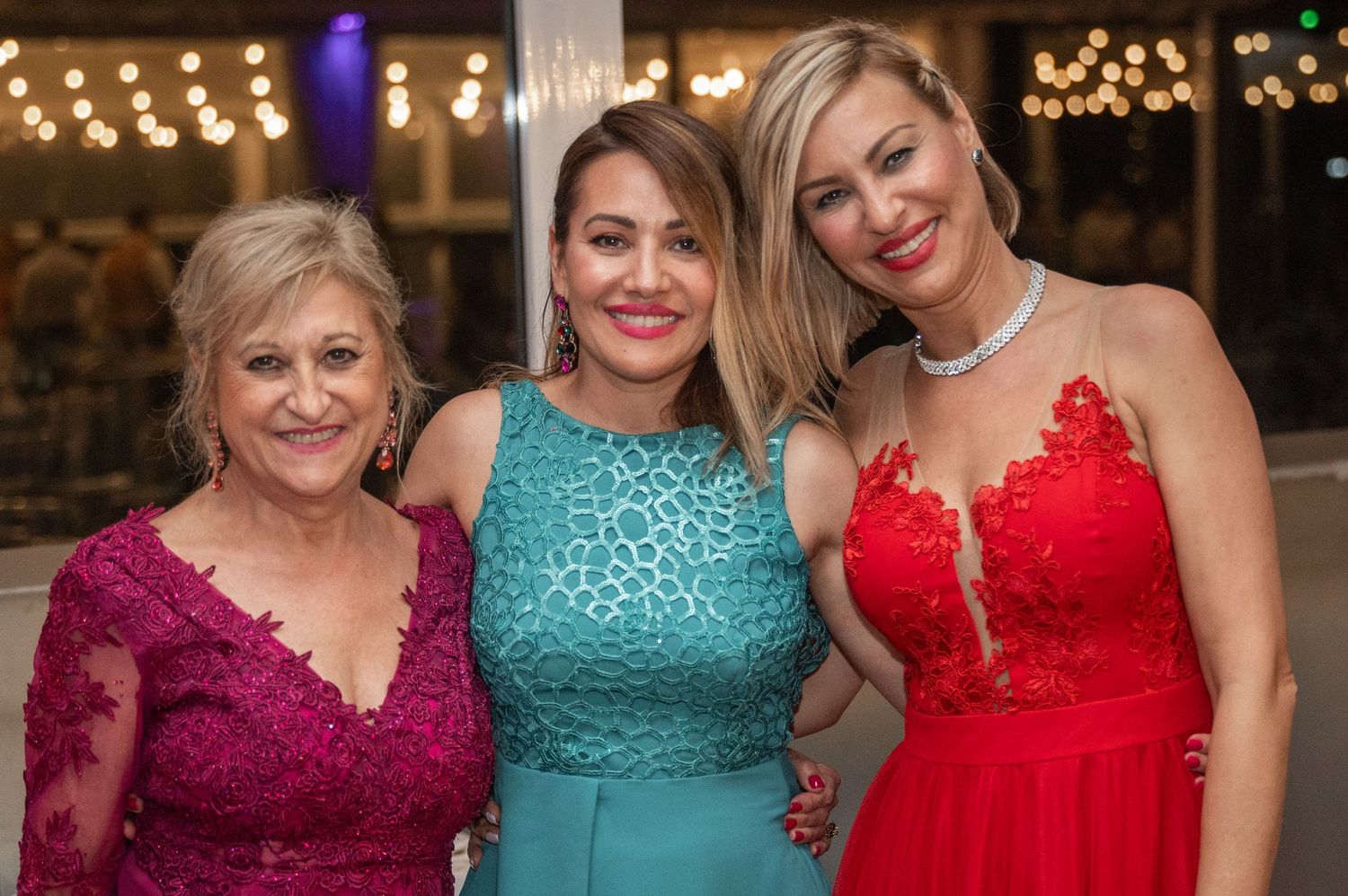 Three women in formal dresses pose together at an evening event with sparkly lights in the background.