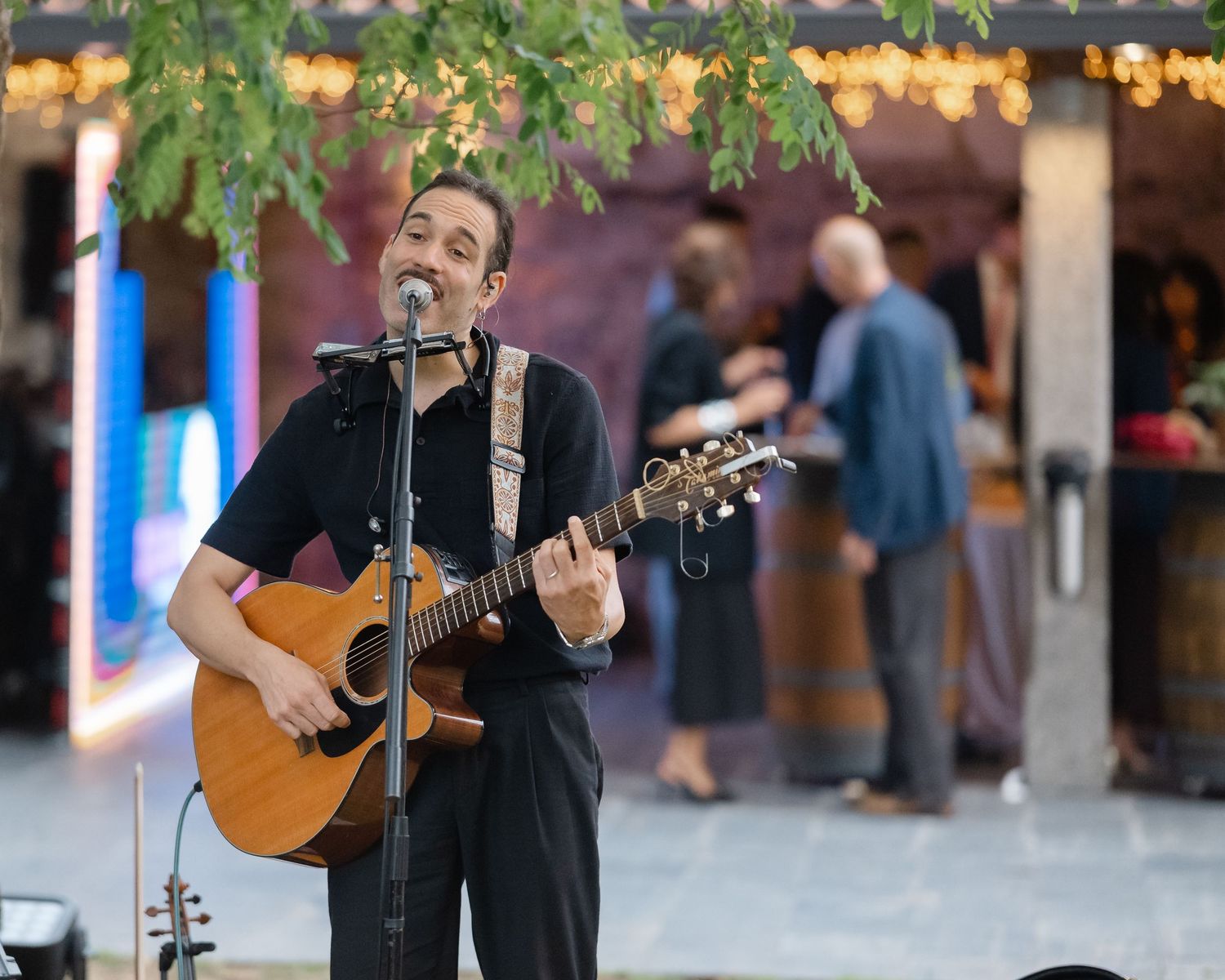 A musician performs with an acoustic guitar on an outdoor stage with string lights and foliage in the background.
