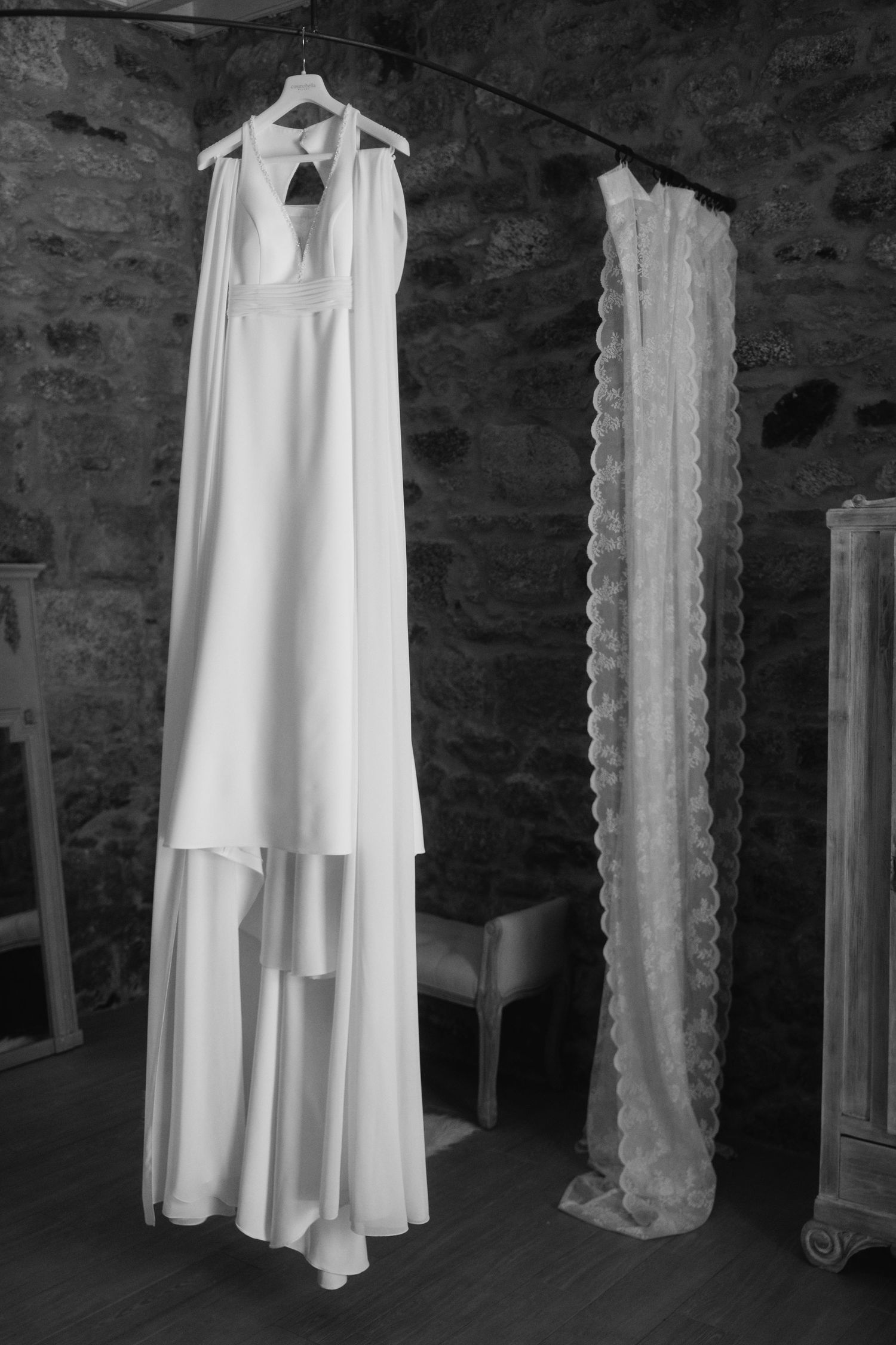 A white wedding dress hangs elegantly against a rustic stone wall with architectural columns.