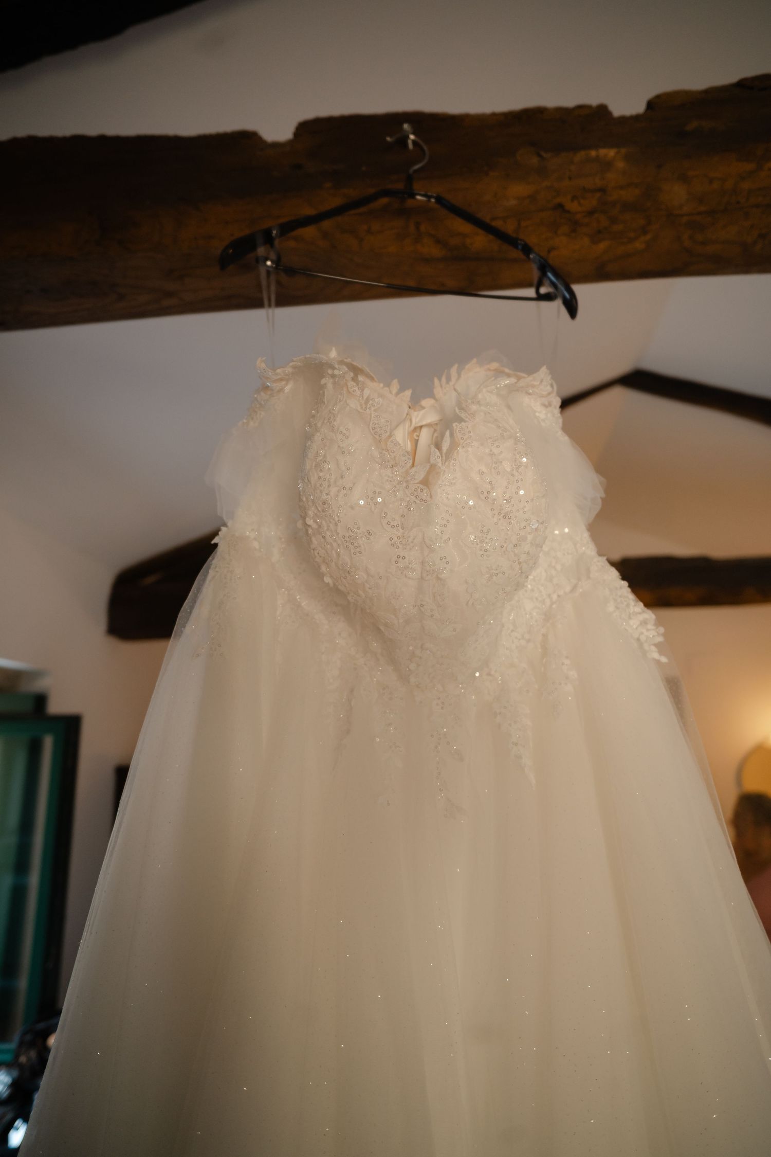 A white wedding dress with delicate lace details hangs from a rustic wooden beam in a room with soft lighting.