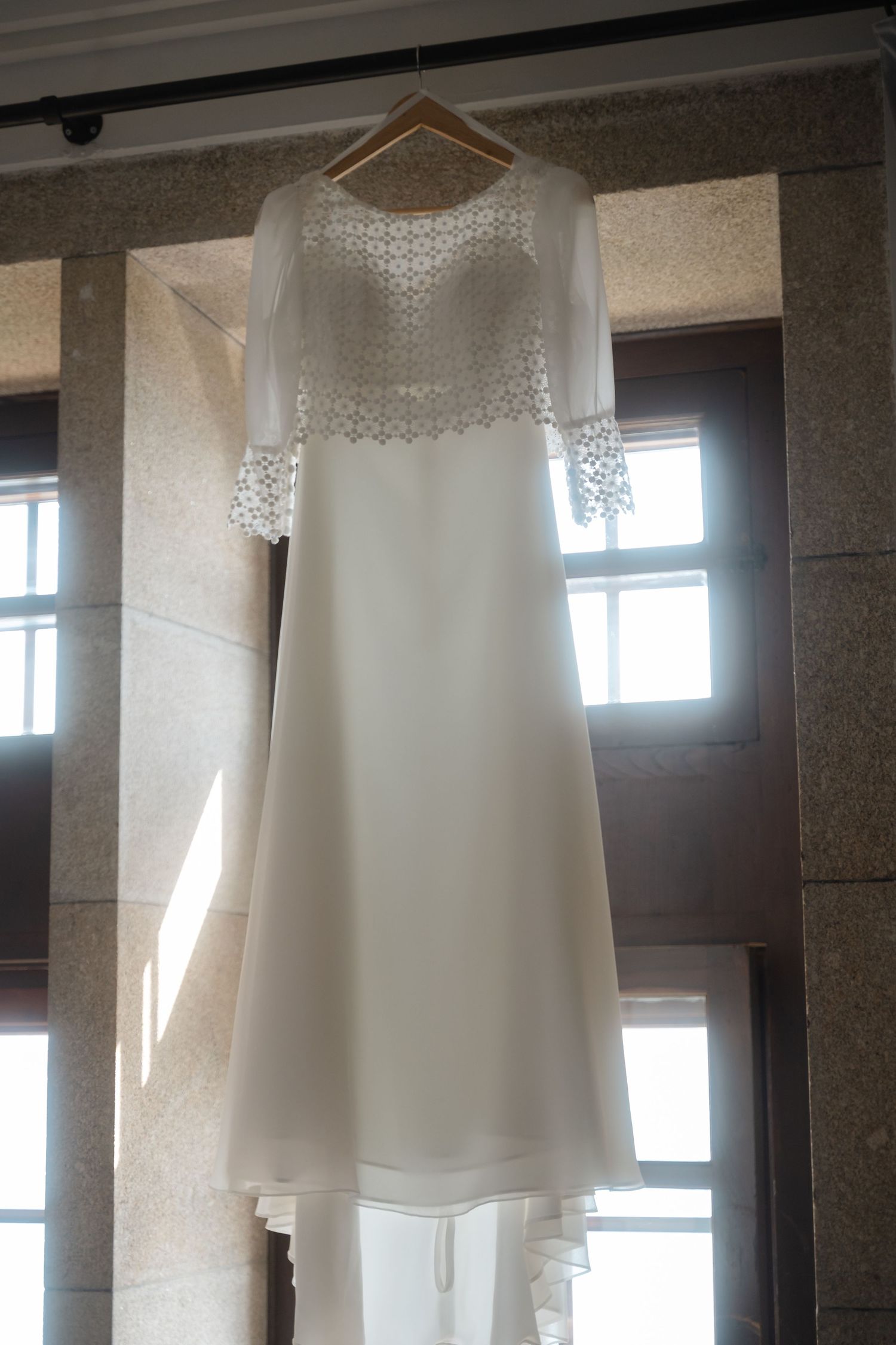 An elegant white wedding dress with delicate lace details hangs in front of sunlit windows.