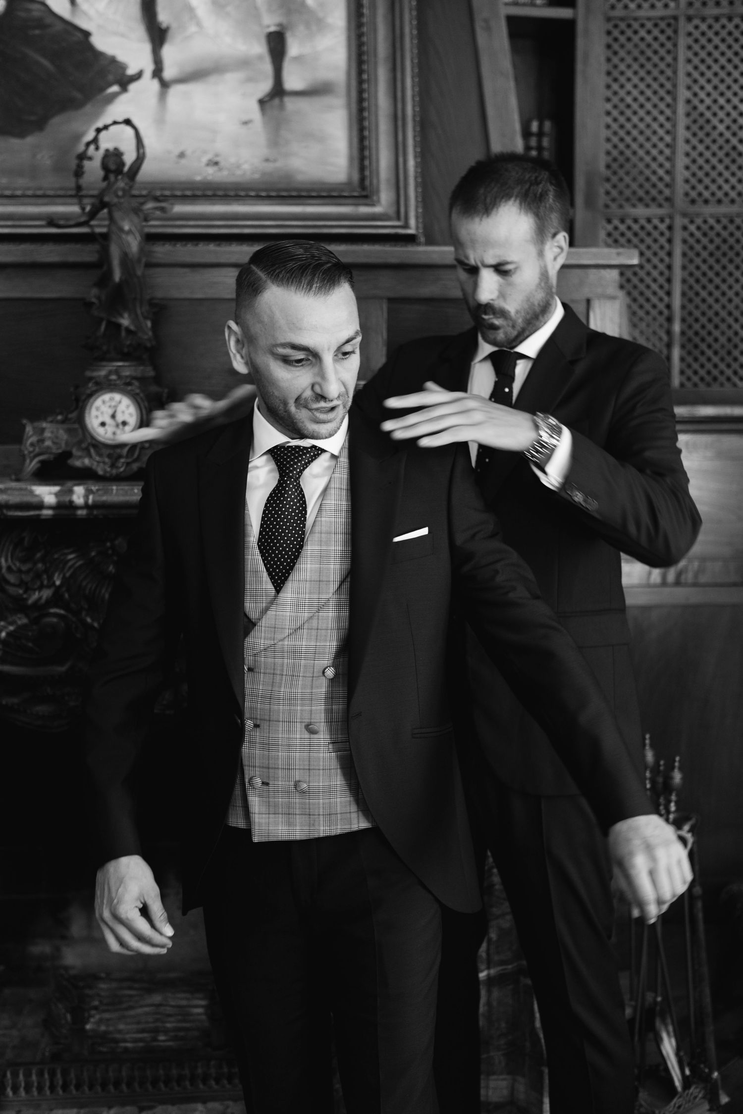 Two men in formal suits making final adjustments to wedding attire in an ornate interior setting.