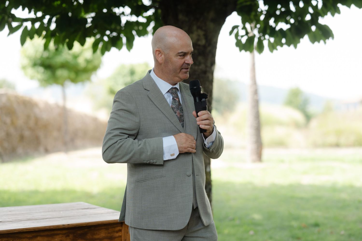 A person in a gray suit holds a microphone while speaking outdoors under a tree on a sunny day.