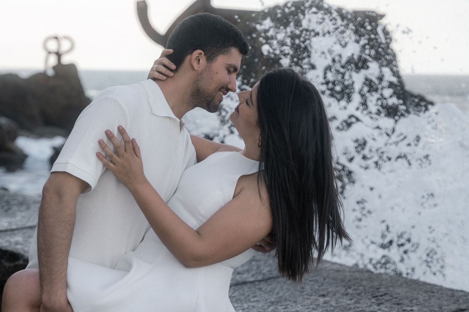 A romantic couple embraces on a rocky beach with crashing waves and ocean spray in the background.