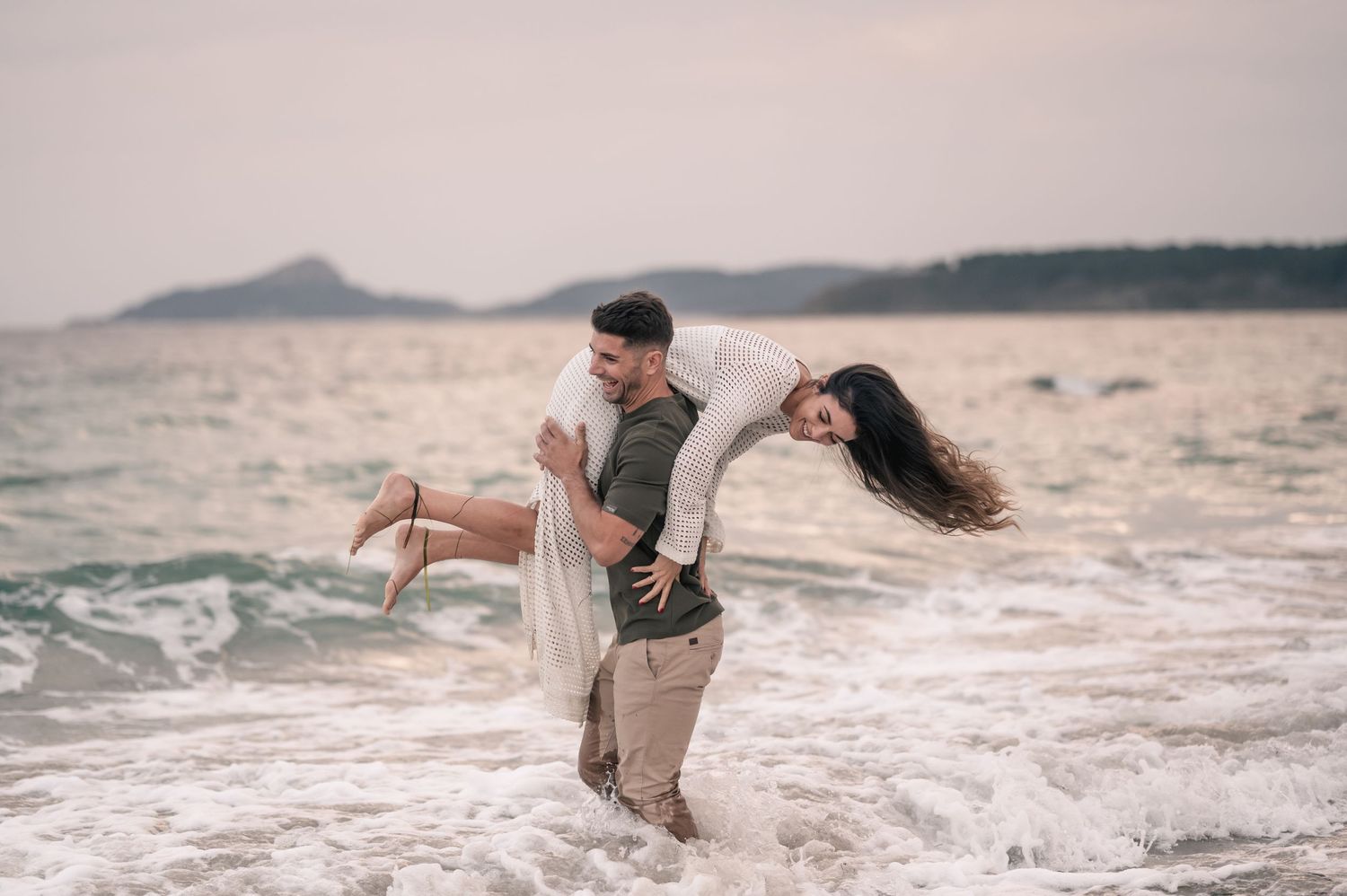 Couple plays joyfully in ocean waves at sunset, creating silhouettes against the water.