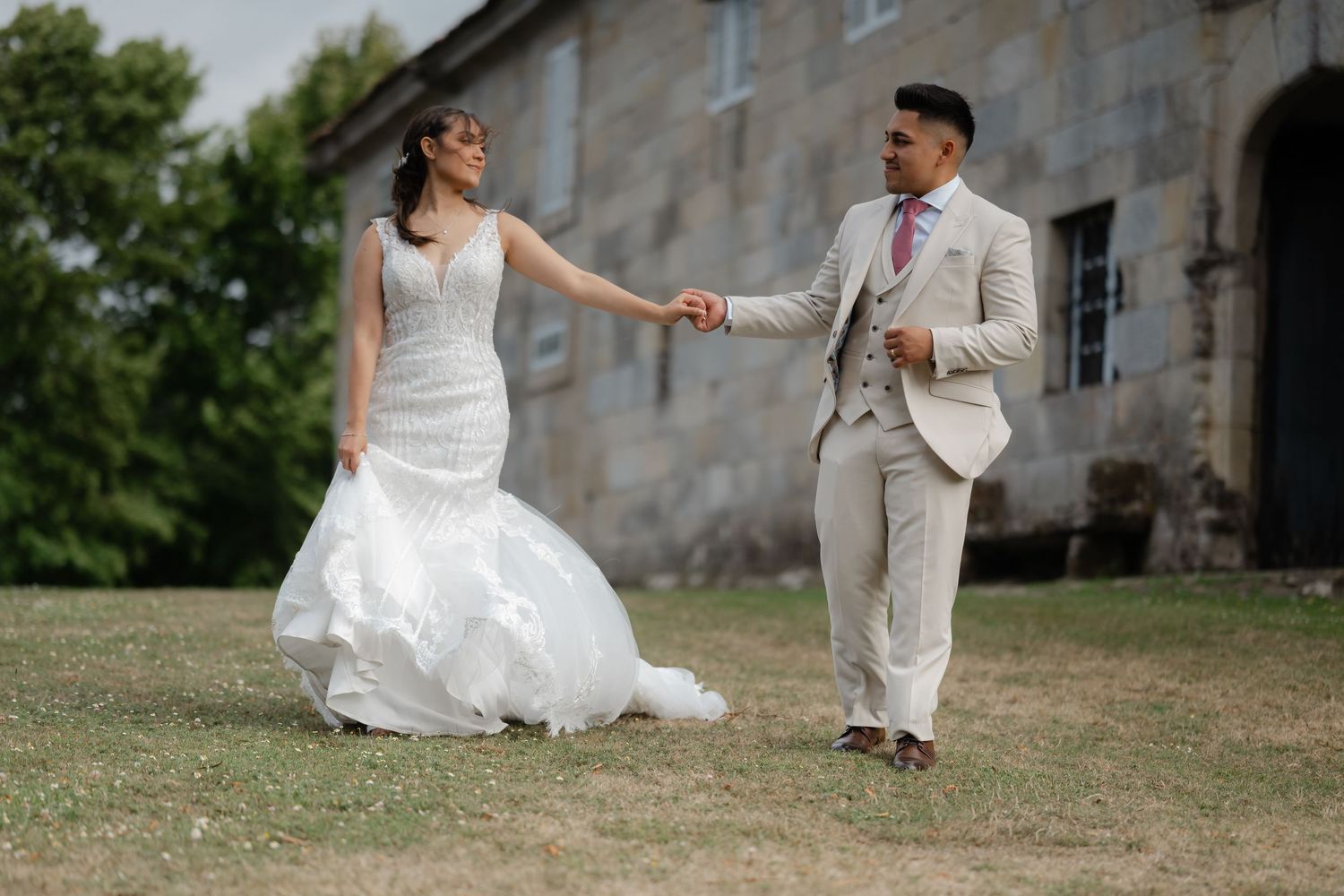 A couple in wedding attire poses together in front of a rustic stone building during their countryside celebration.