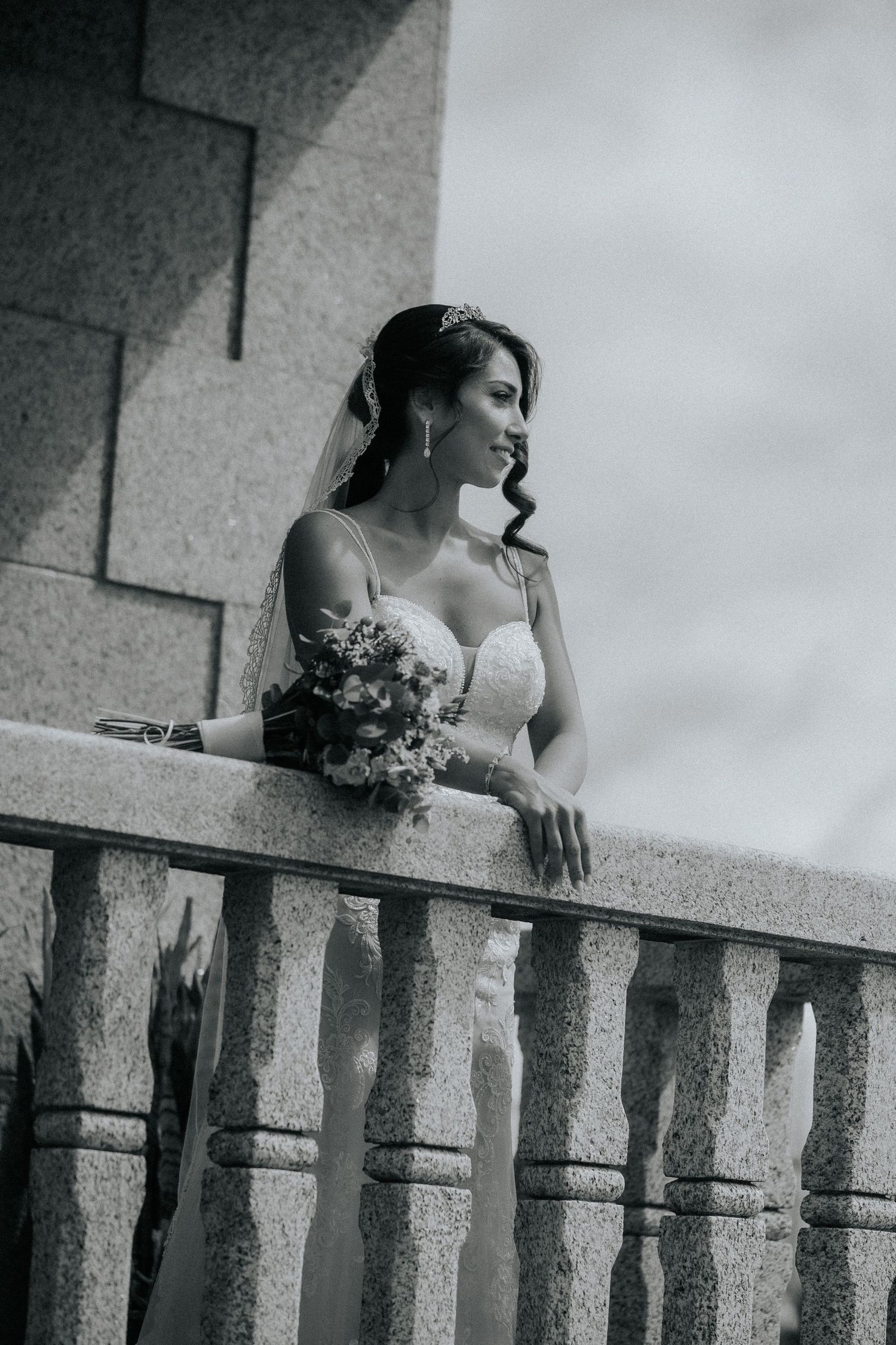 Black and white photo of a bride leaning on a stone balcony with a bouquet.