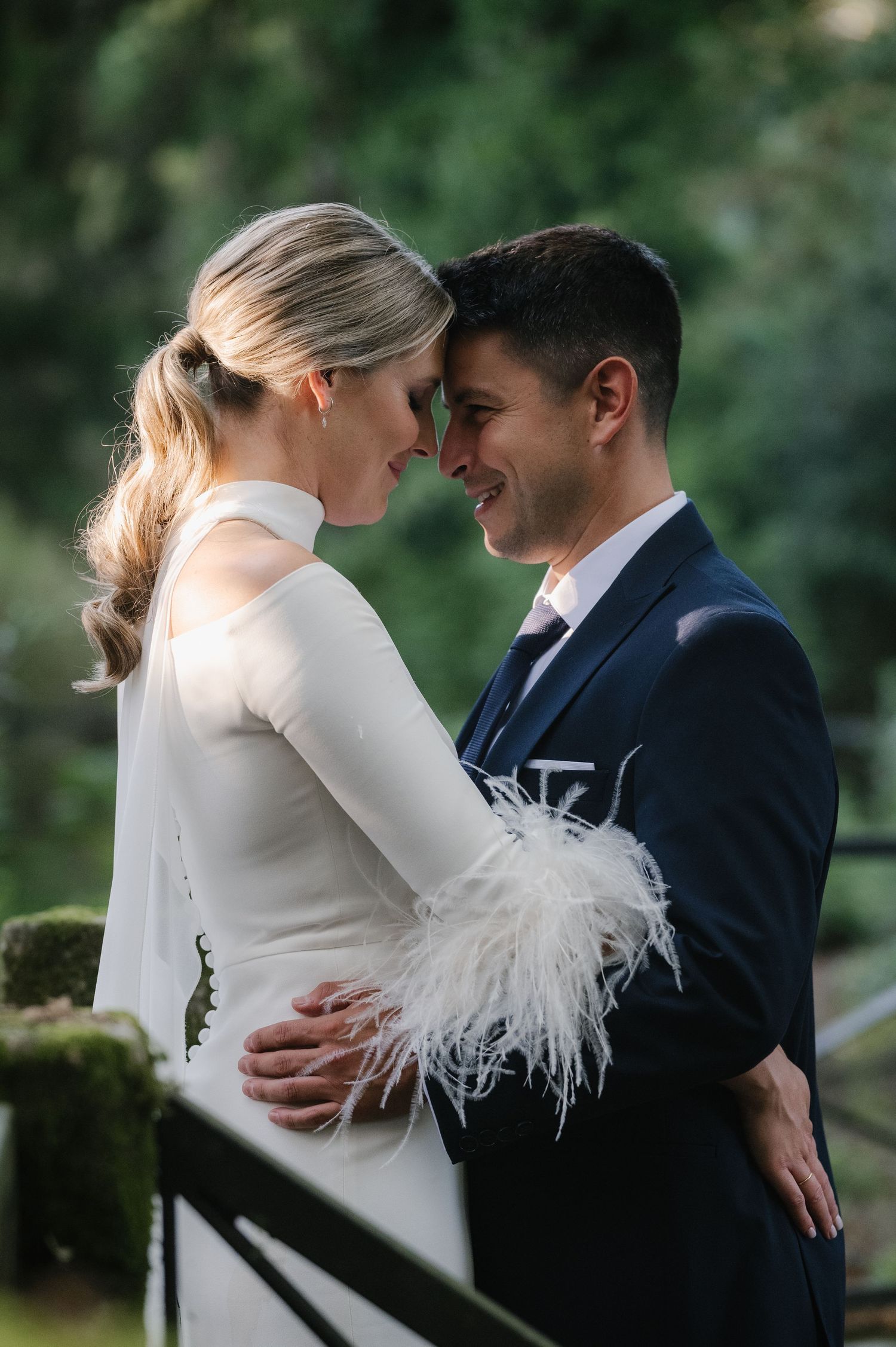 Romantic wedding couple embracing in a garden with feathered sleeve detail on bride's white gown.
