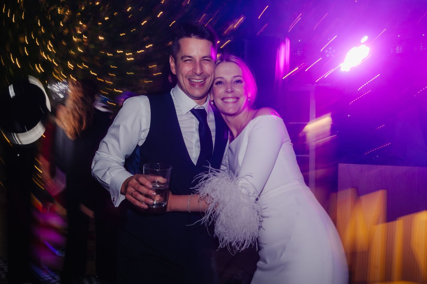 People dancing and celebrating at a wedding reception with purple lighting and festive atmosphere.