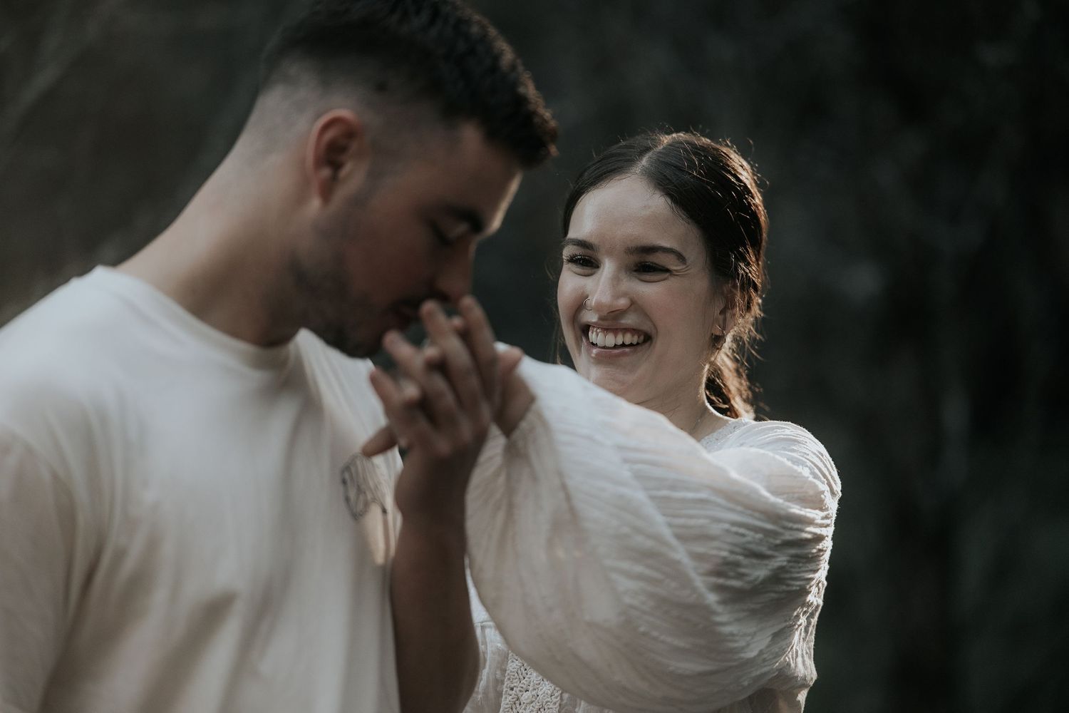 An intimate couple shares tender moments in white clothing against a dark forest backdrop.