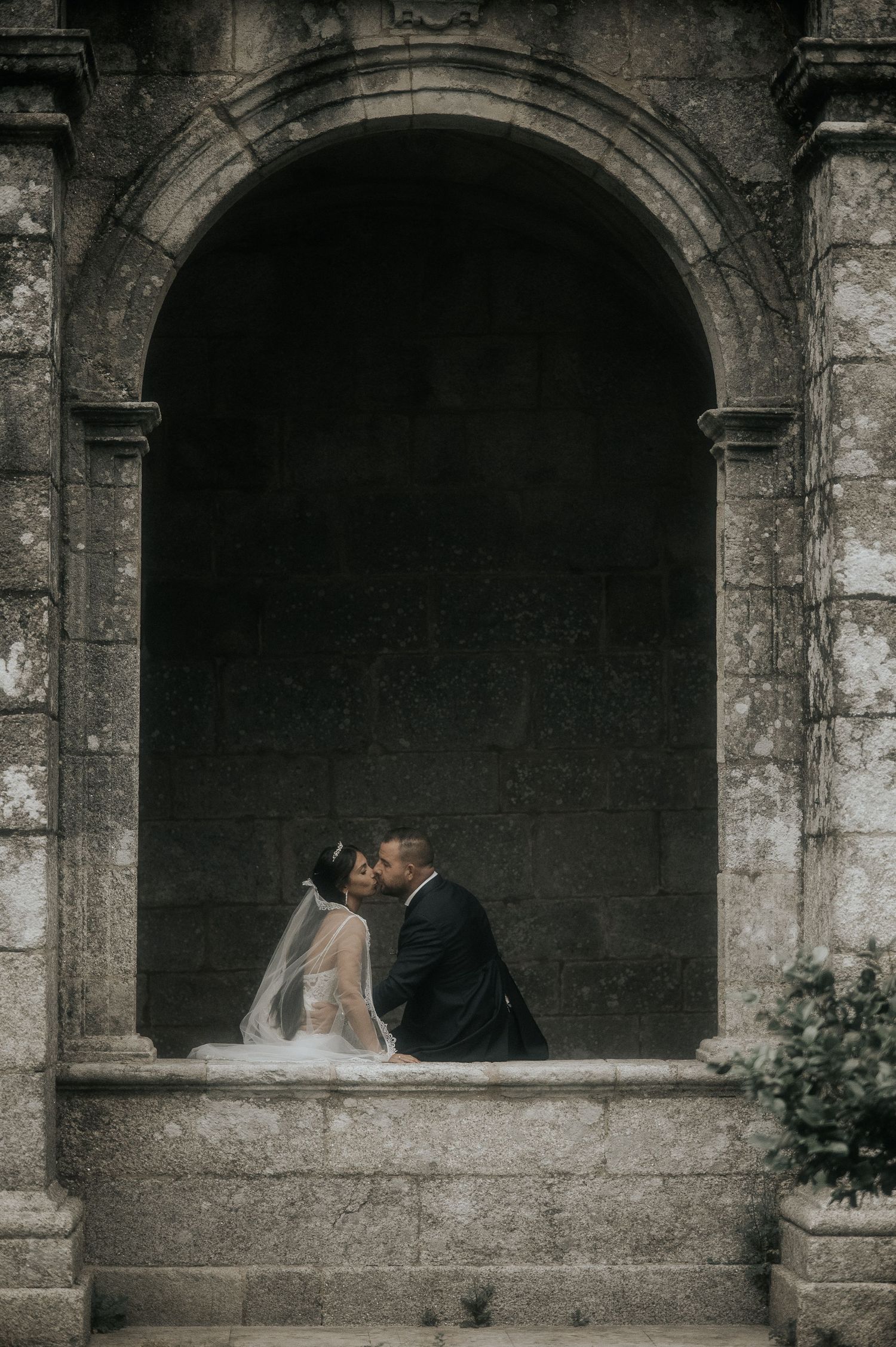A romantic wedding couple embraces in a stone archway of an ancient building with dramatic moody lighting.