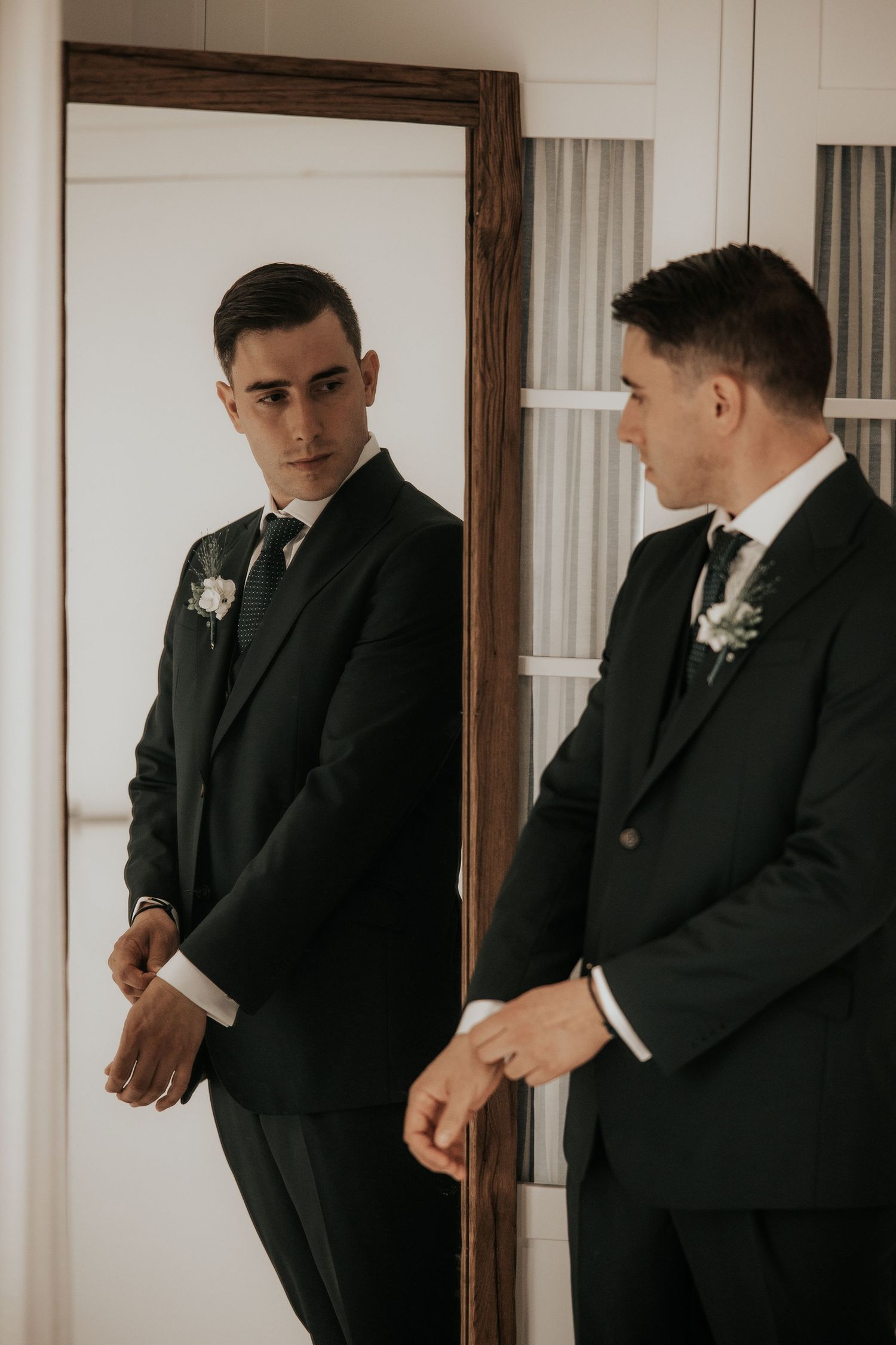 A groom in a classic black suit with boutonniere looks at his reflection in a mirror while getting ready.