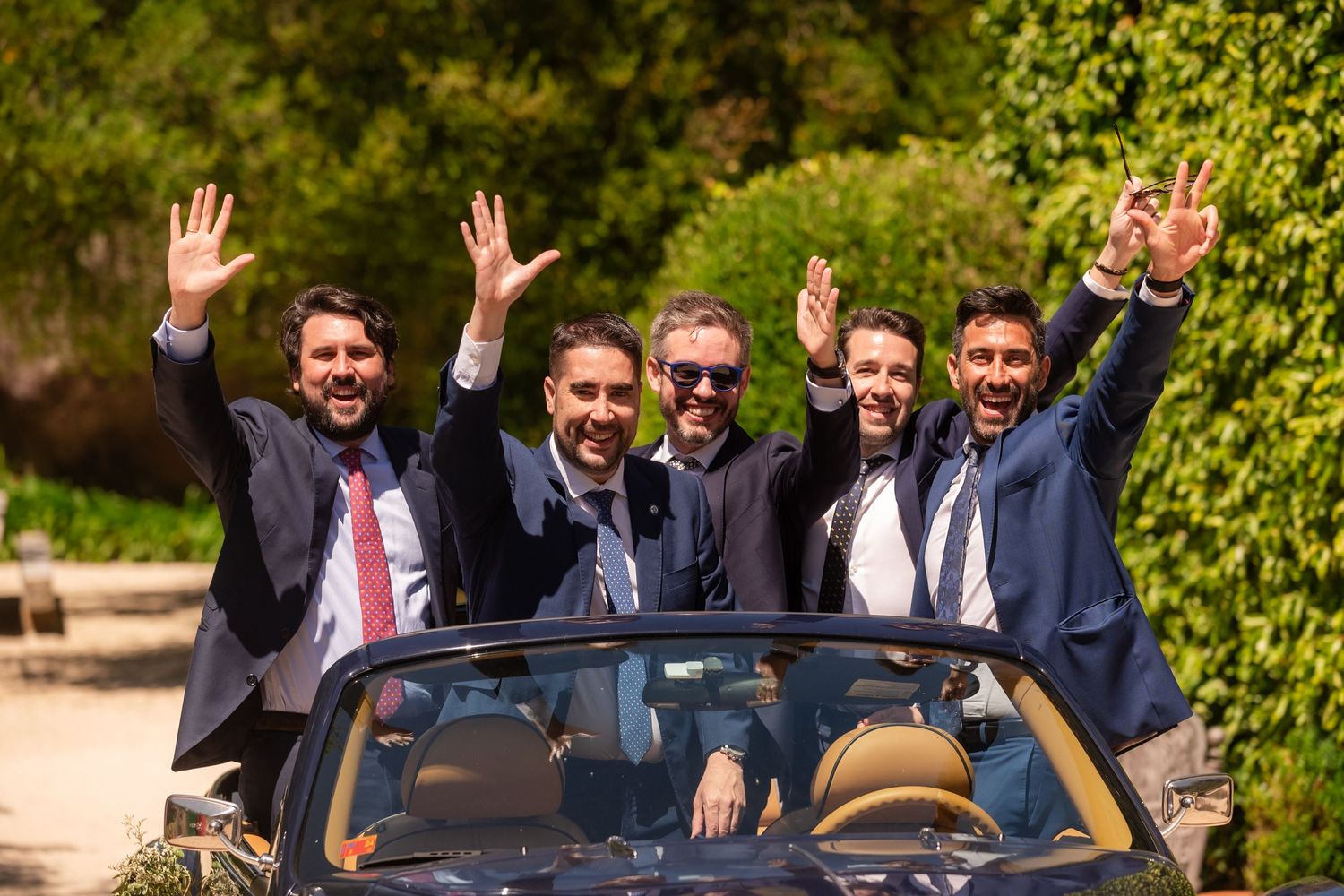 A group of cheerful people in formal attire wave while riding in a convertible car with greenery in the background.