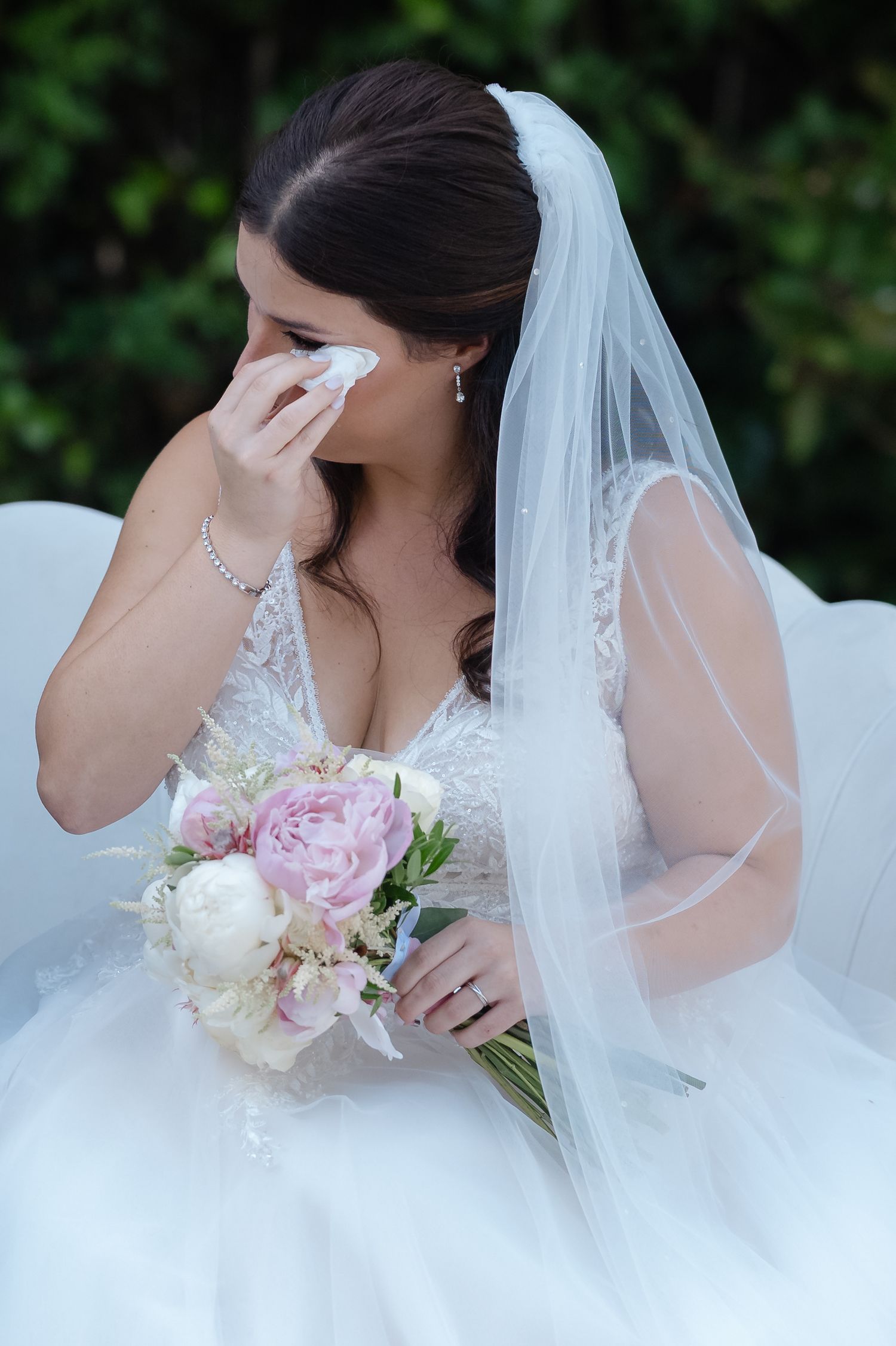 A bride in a white wedding dress holds a pink and white bouquet while wearing a delicate veil in an outdoor setting.