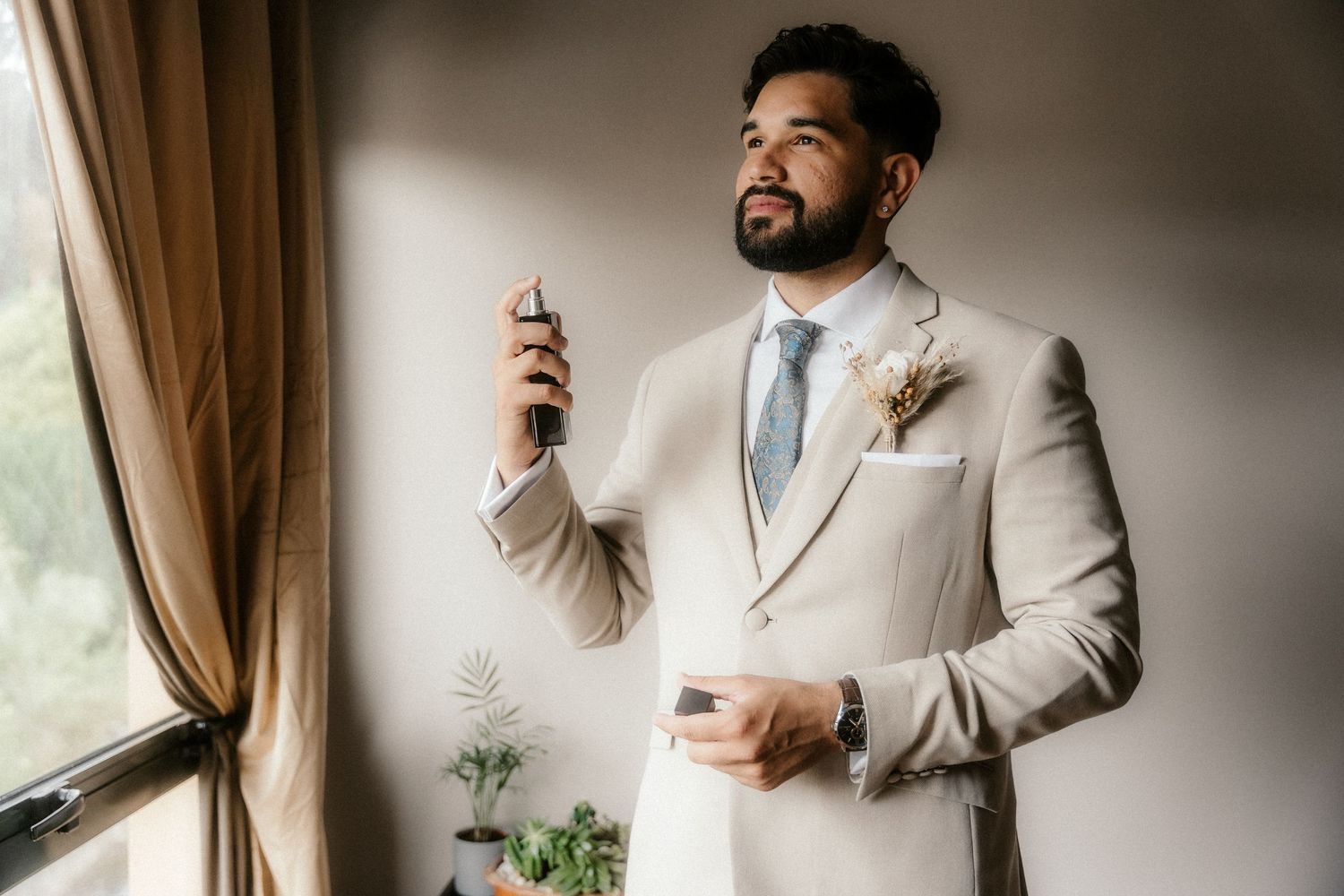 A groom in a light beige suit adjusts his tie while standing near a window in an elegant room.