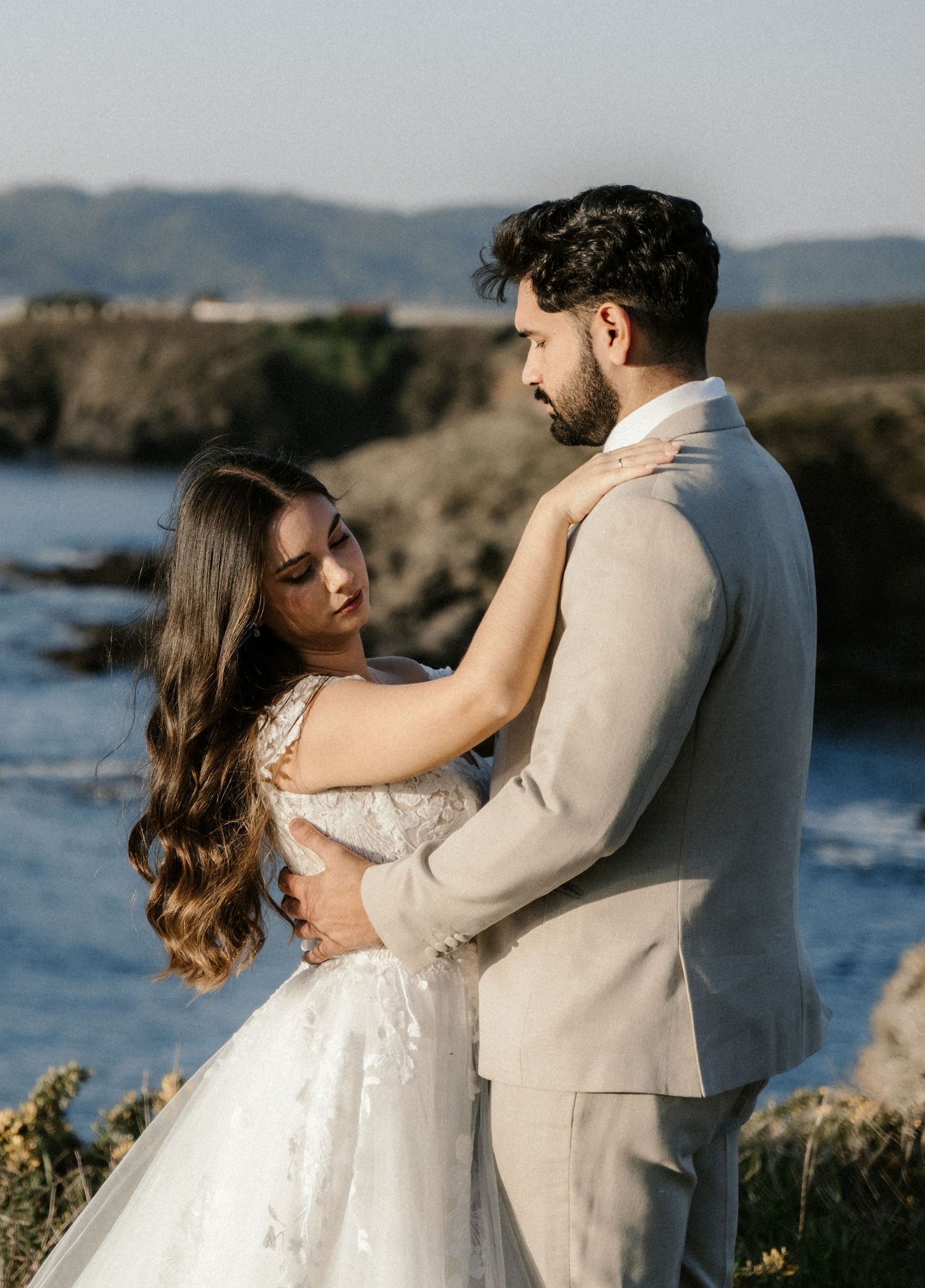 A couple shares a romantic moment in their wedding attire along a scenic coastal cliff.