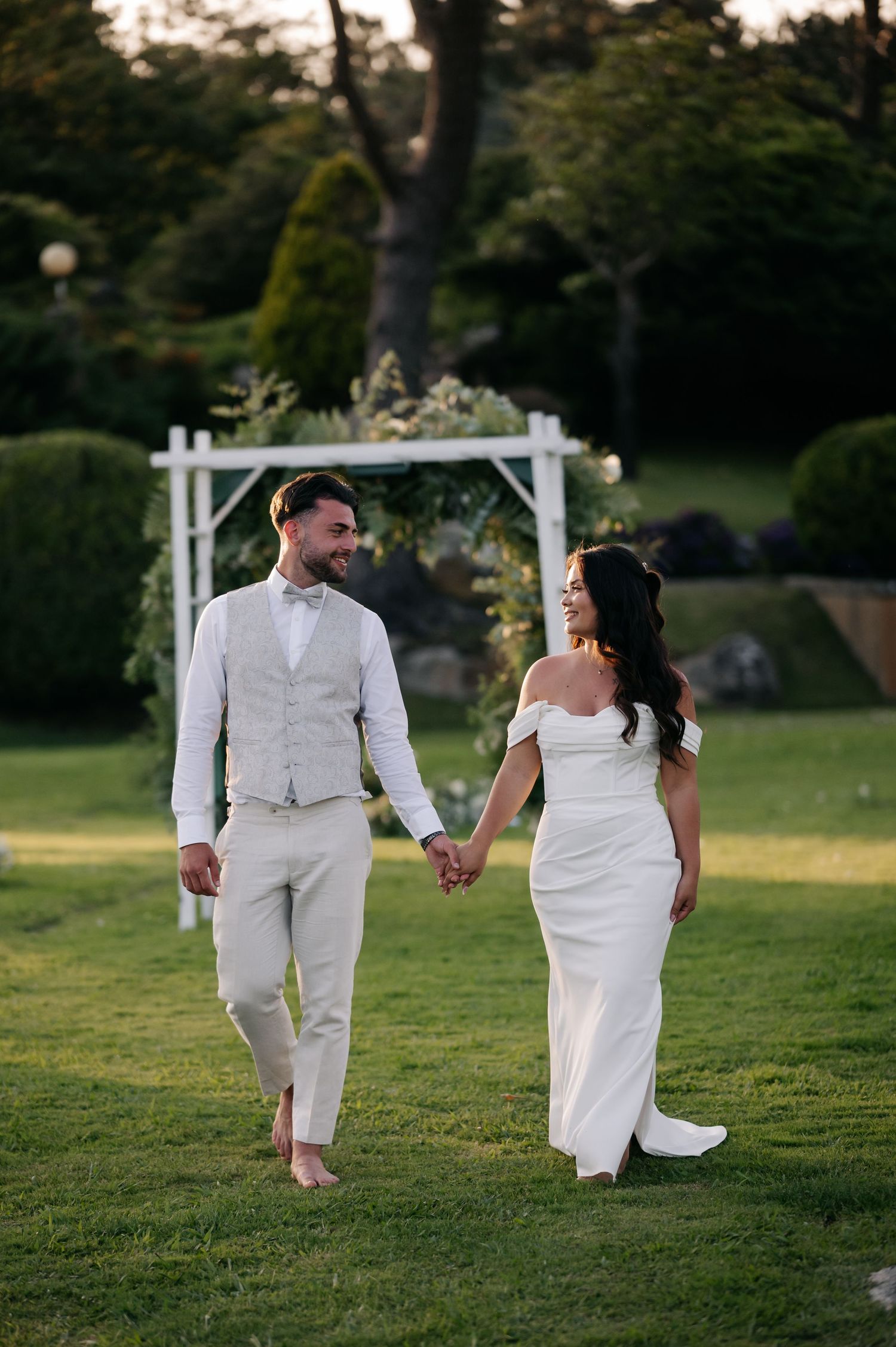 A couple in wedding attire walks hand in hand across a lush green lawn during sunset.