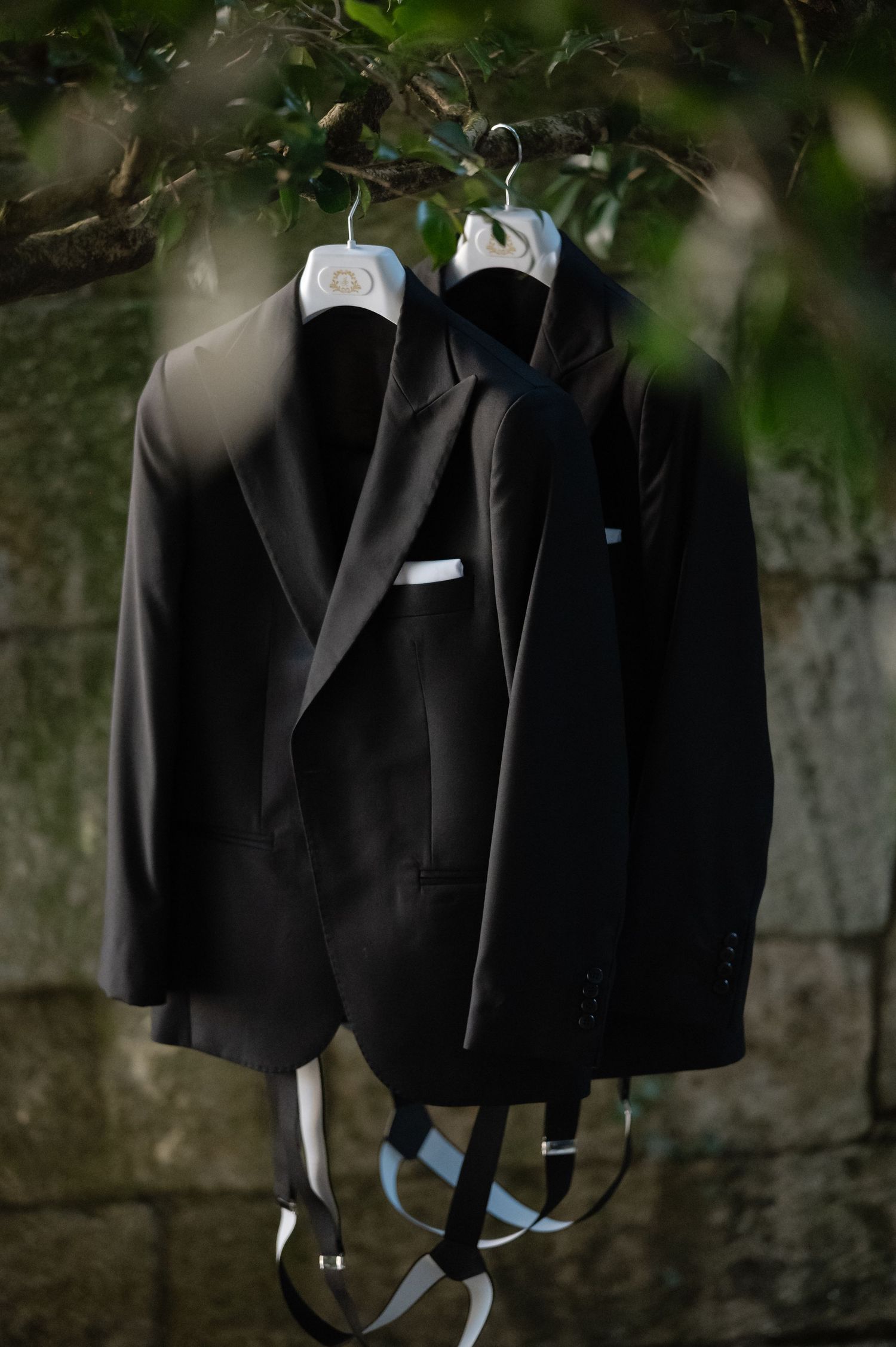 Black suit jackets hanging on wooden hangers against a stone wall with natural lighting.