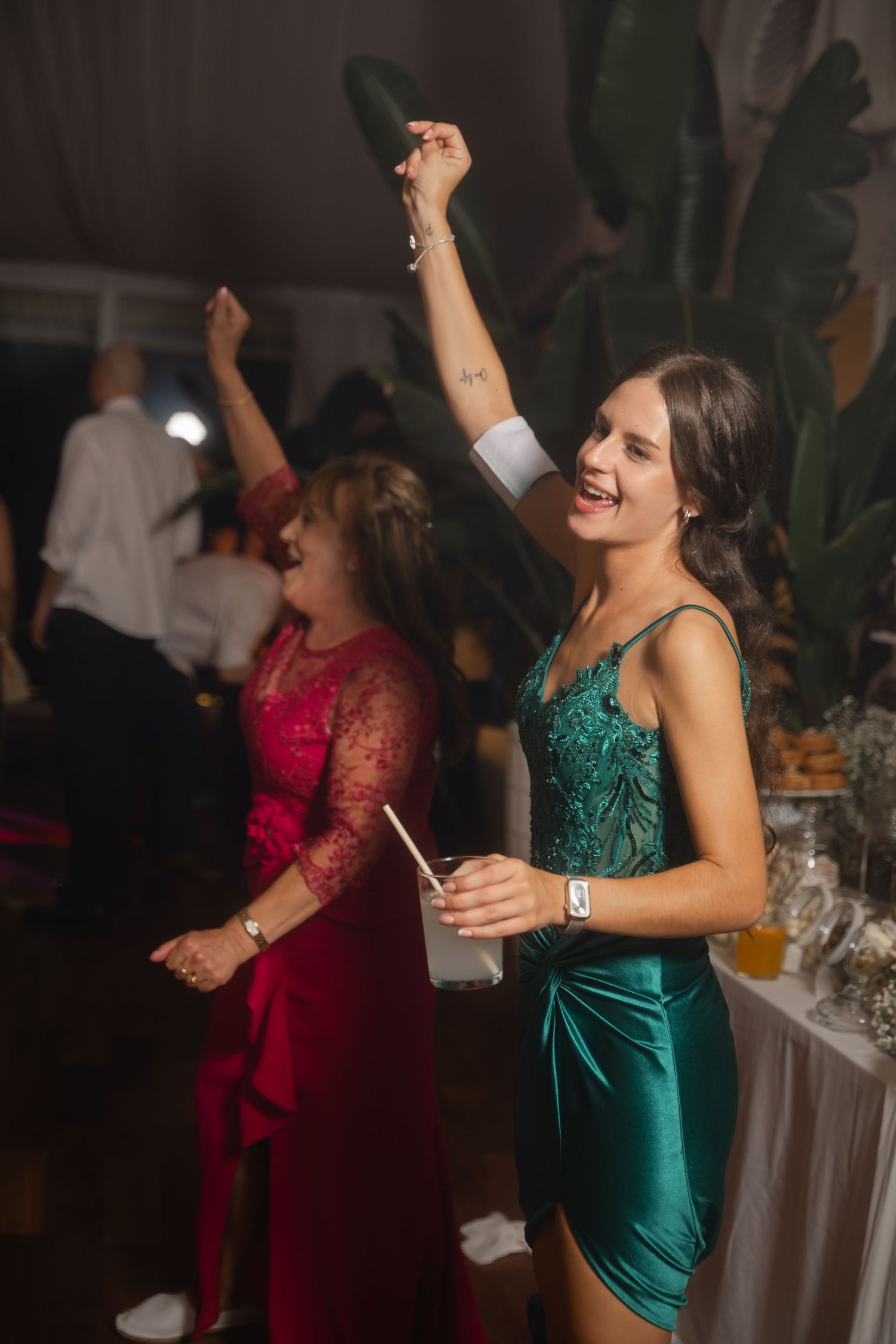 Two party guests in elegant dresses dance energetically on a dimly lit dance floor near tropical plants.