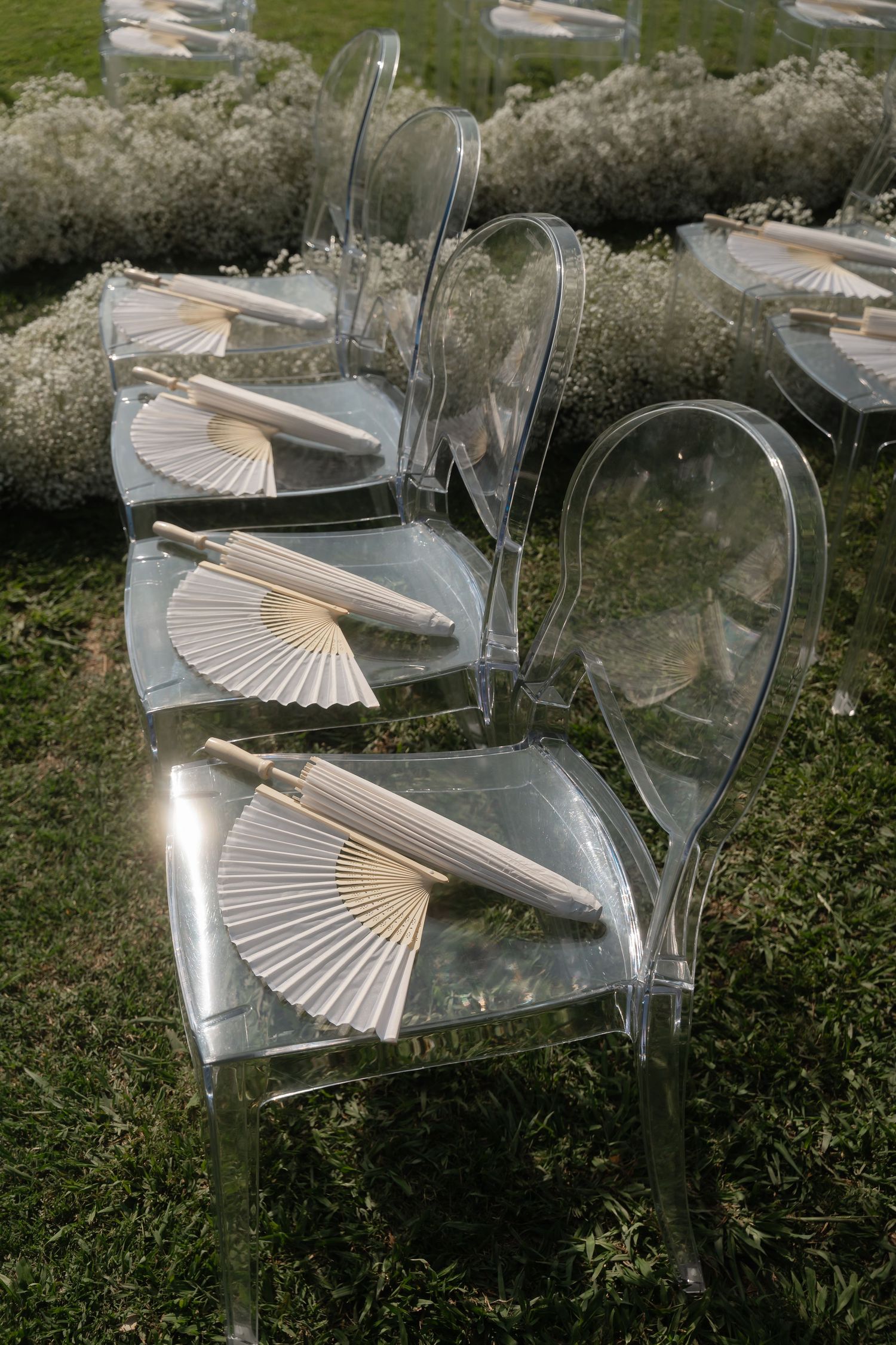 Clear acrylic ghost chairs decorated with white paper fans for an elegant outdoor wedding ceremony setup.